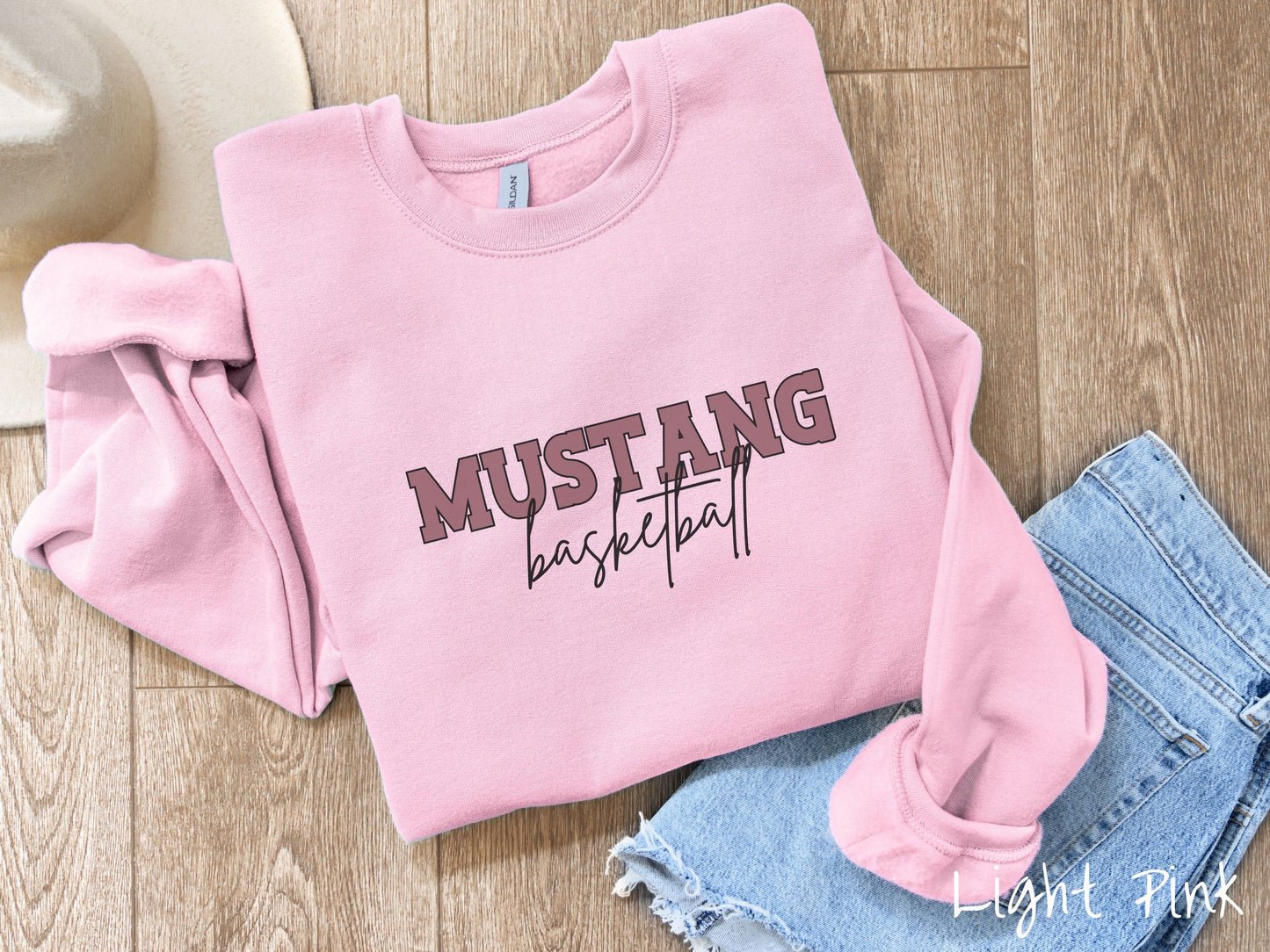A cute, vintage light pink colored sweatshirt with the word Mustang in uppercase, purple font across the front and the word basketball below that in black, undercase cursive writing.