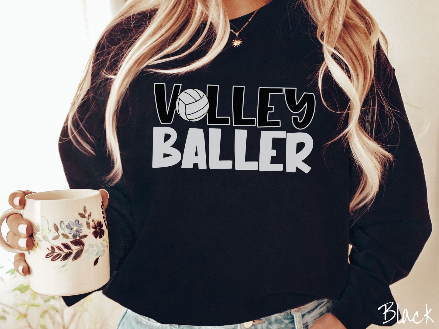 A woman wearing a cute, vintage black colored sweatshirt with the word Volley across the front in black, uppercase font. The O in Volley is a white volleyball. Below that is the word Baller in white, uppercase font.