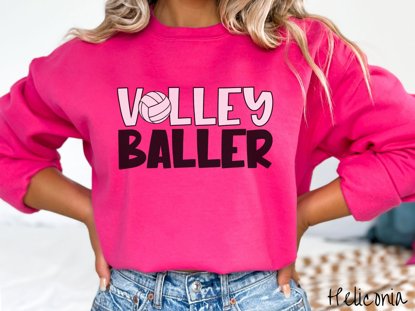 A woman wearing a cute, vintage heliconia colored sweatshirt with the word Volley across the front in white, uppercase font. The O in Volley is a white volleyball. Below that is the word Baller in black, uppercase font.
