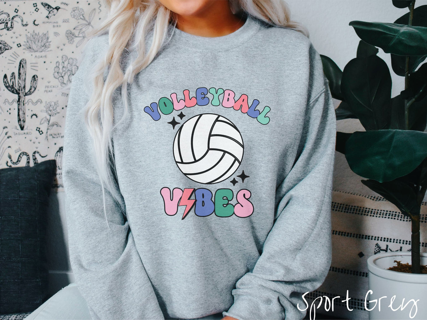 A woman wearing a cute, vintage sport grey colored sweatshirt with the word Volleyball across the front in colorful, uppercase fonts. Below that is a white volleyball and below that is the word vibes in colorful, uppercase fonts.