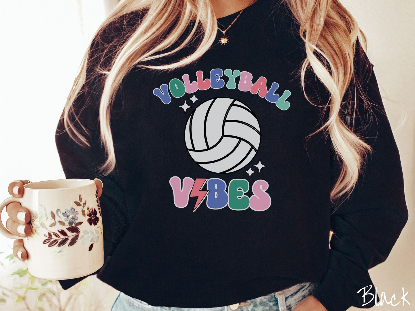 A woman wearing a cute, vintage black colored sweatshirt with the word Volleyball across the front in colorful, uppercase fonts. Below that is a white volleyball and below that is the word vibes in colorful, uppercase fonts.