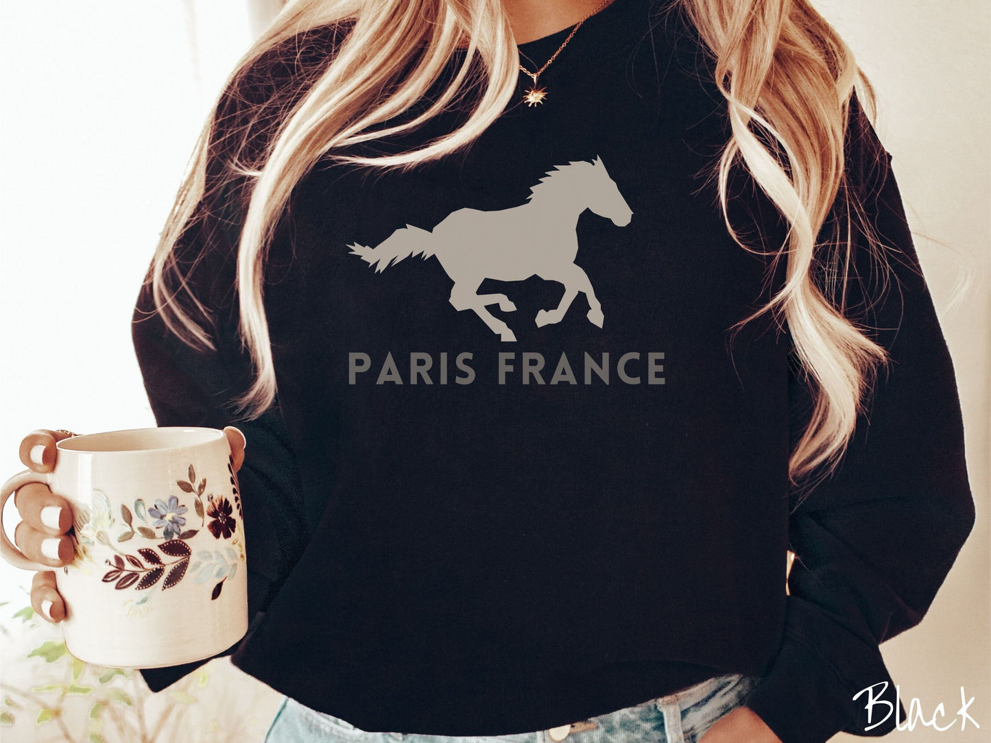 A woman wearing a cute, vintage black colored comfy sweatshirt with a gray silhouette of a running horse across the font. Below it are the words Paris France in gray, uppercase font.