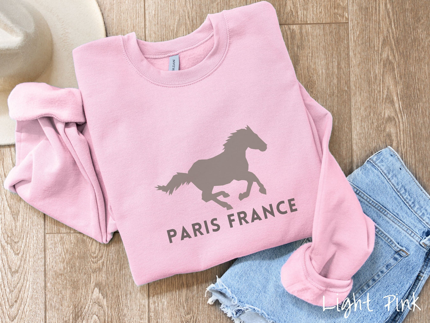 A cute, vintage light pink colored comfy sweatshirt with a gray silhouette of a running horse across the font. Below it are the words Paris France in gray, uppercase font.