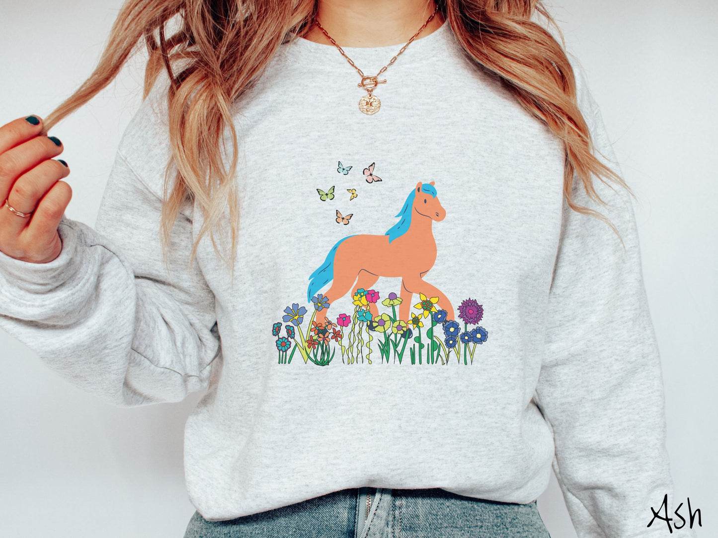 A woman wearing a cute, vintage ash colored comfy sweatshirt with a brown horse with blue hair walking through a field of colorful flowers and grass. There are colorful butterflies flying above the horse.