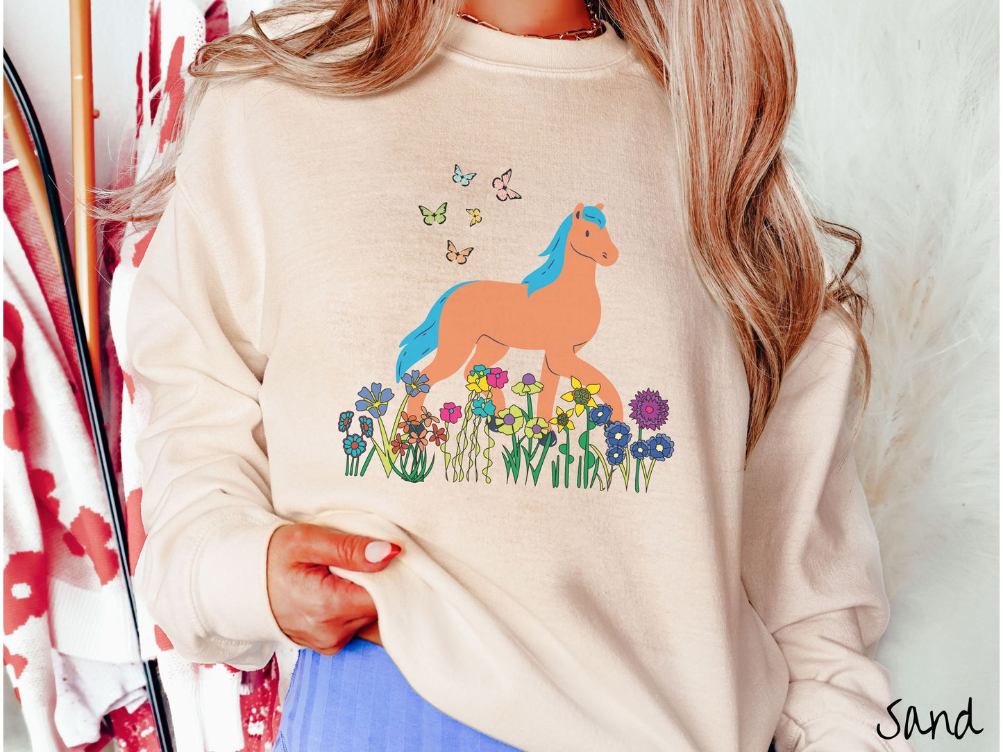 A woman wearing a cute, vintage sand colored comfy sweatshirt with a brown horse with blue hair walking through a field of colorful flowers and grass. There are colorful butterflies flying above the horse.