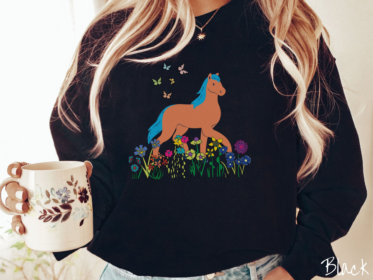 A woman wearing a cute, vintage black colored comfy sweatshirt with a brown horse with blue hair walking through a field of colorful flowers and grass. There are colorful butterflies flying above the horse.