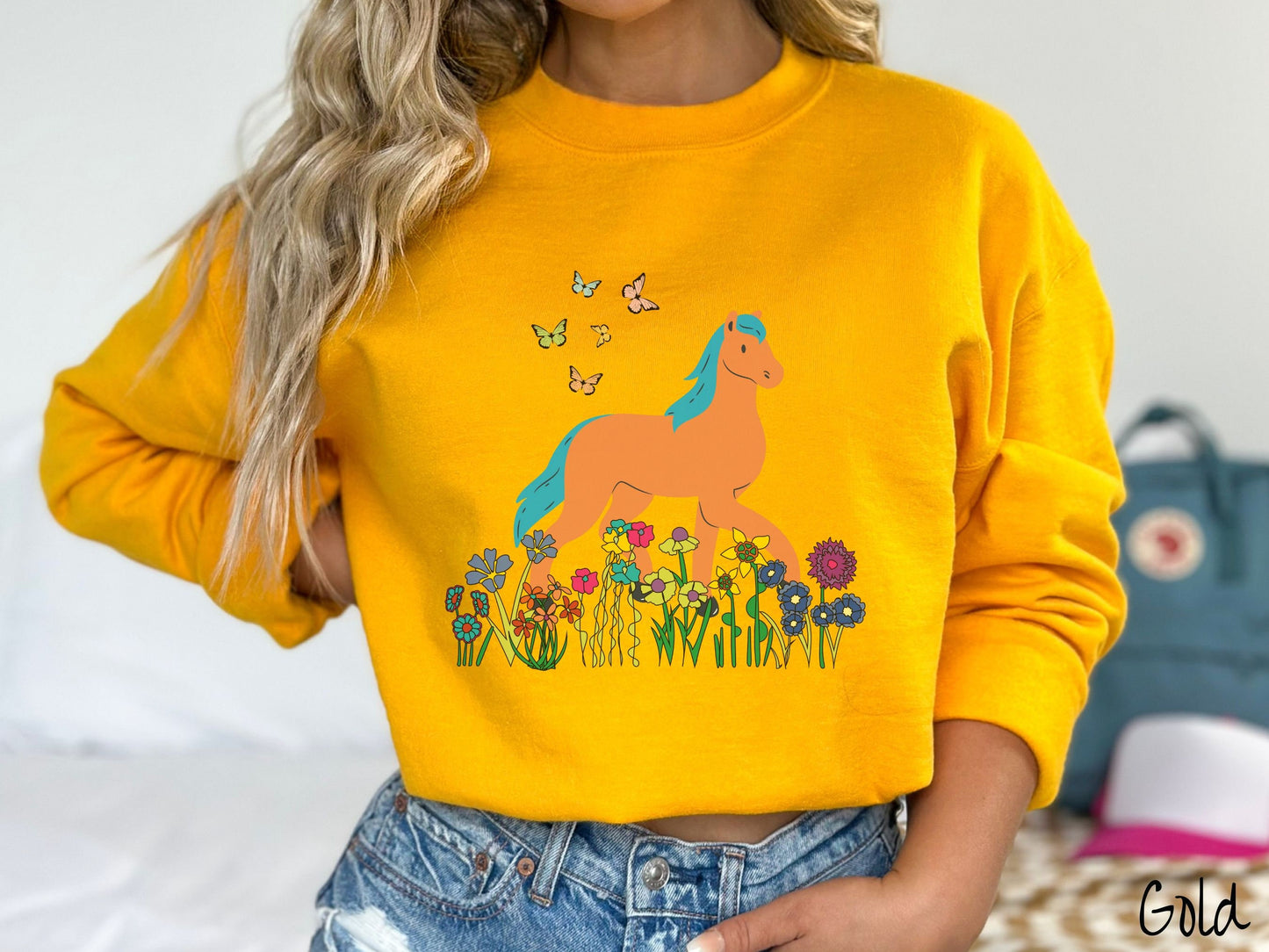 A woman wearing a cute, vintage gold colored comfy sweatshirt with a brown horse with blue hair walking through a field of colorful flowers and grass. There are colorful butterflies flying above the horse.