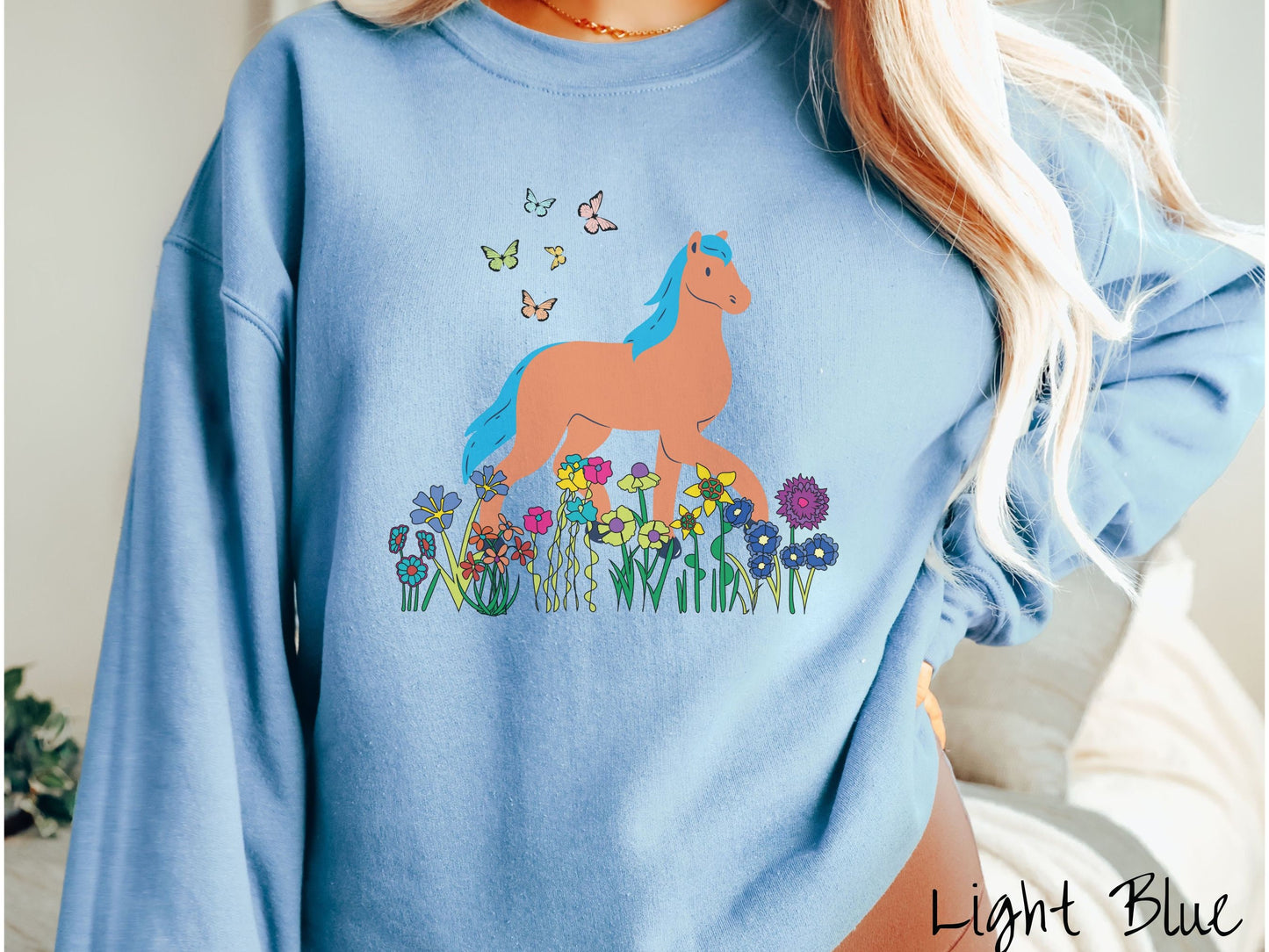 A woman wearing a cute, vintage light blue colored comfy sweatshirt with a brown horse with blue hair walking through a field of colorful flowers and grass. There are colorful butterflies flying above the horse.