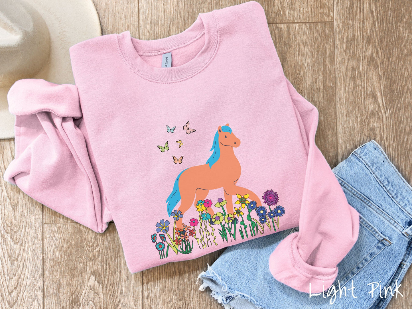 A cute, vintage light pink colored comfy sweatshirt with a brown horse with blue hair walking through a field of colorful flowers and grass. There are colorful butterflies flying above the horse.