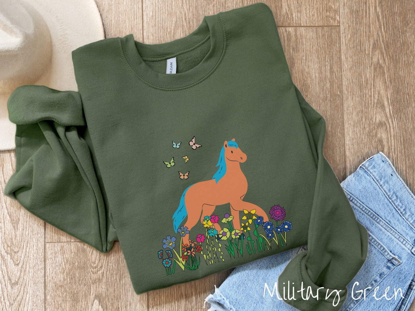 A woman wearing a cute, vintage miltary green colored comfy sweatshirt with a brown horse with blue hair walking through a field of colorful flowers and grass. There are colorful butterflies flying above the horse.