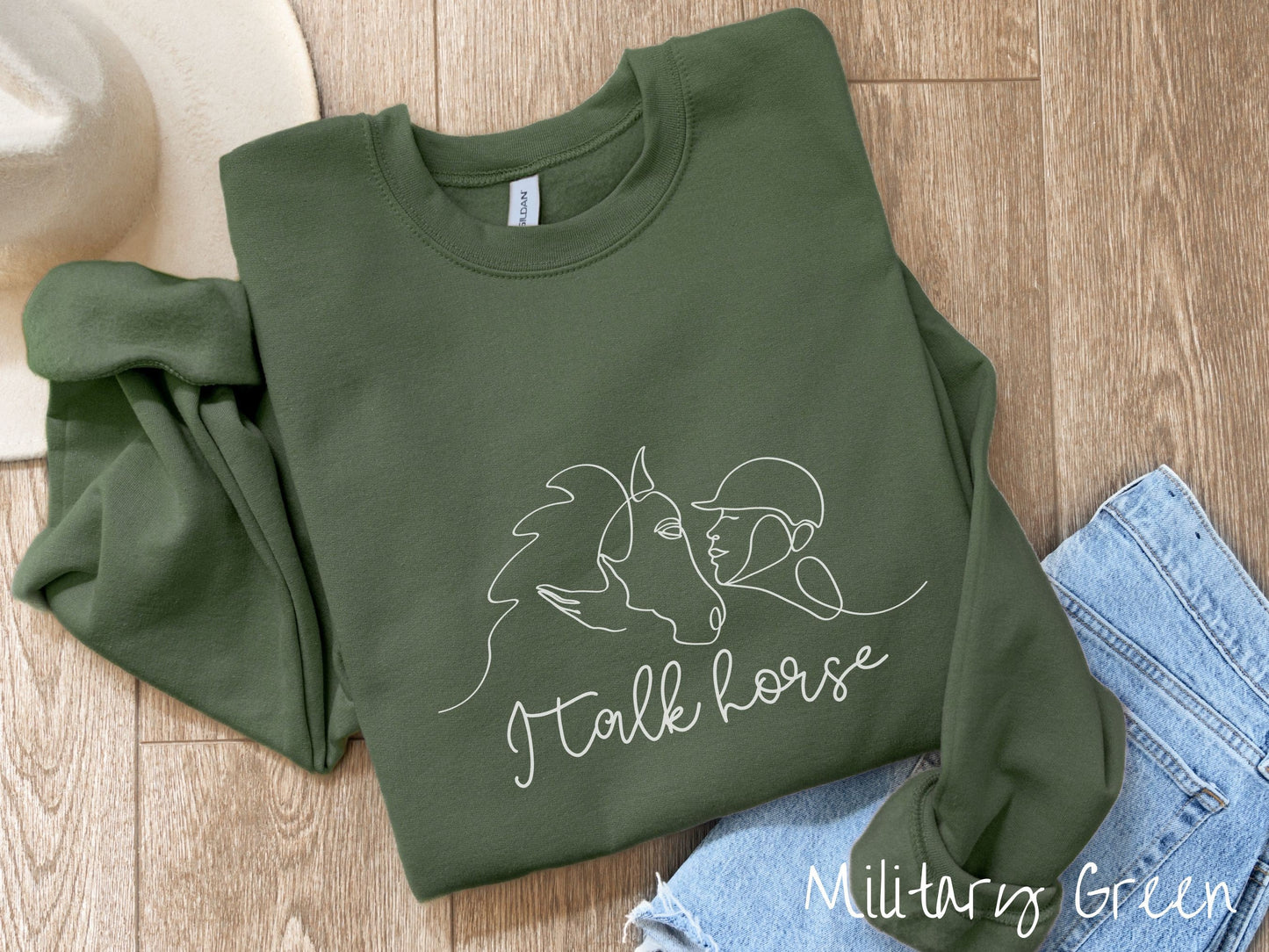 A woman wearing a cute, vintage military green colored comfy sweatshirt with a white silhouette outline of a horse jockey embracing his horse. Below that are the words I talk horse in white, cursive font.