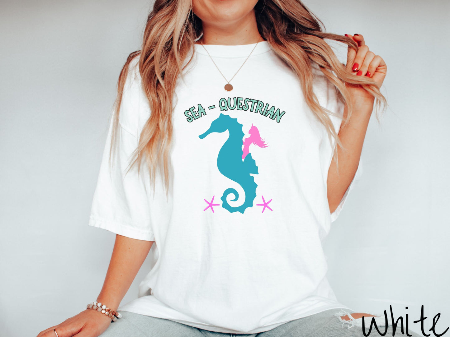 A woman wearing a cute, vintage white colored Comfort Colors shirt with the text Sea-Questrian in seafoam colored font across the top. Below this is a pink mermaid with flowing hair riding on the back of a turquoise seahorse with pink starfish below.