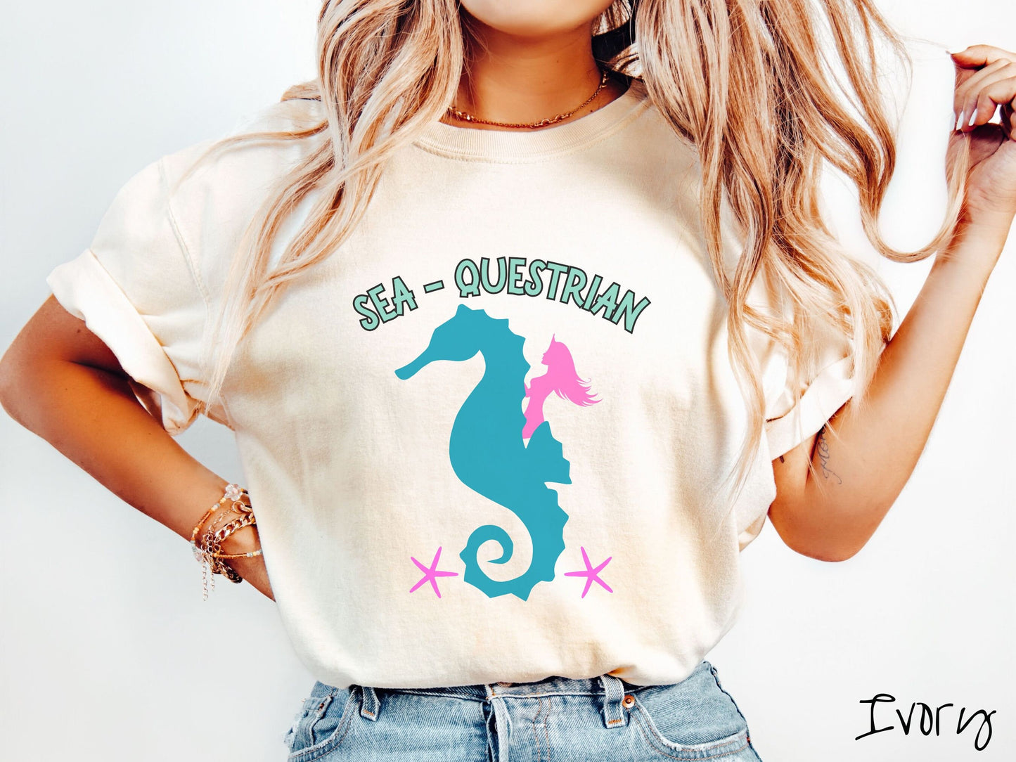 A woman wearing a cute, vintage ivory colored Comfort Colors shirt with the text Sea-Questrian in seafoam colored font across the top. Below this is a pink mermaid with flowing hair riding on the back of a turquoise seahorse with pink starfish below.