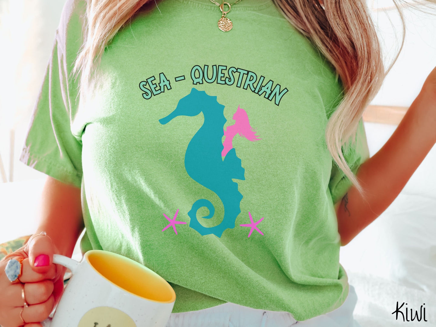 A woman wearing a cute, vintage kiwi colored Comfort Colors shirt with the text Sea-Questrian in seafoam colored font across the top. Below this is a pink mermaid with flowing hair riding on the back of a turquoise seahorse with pink starfish below.