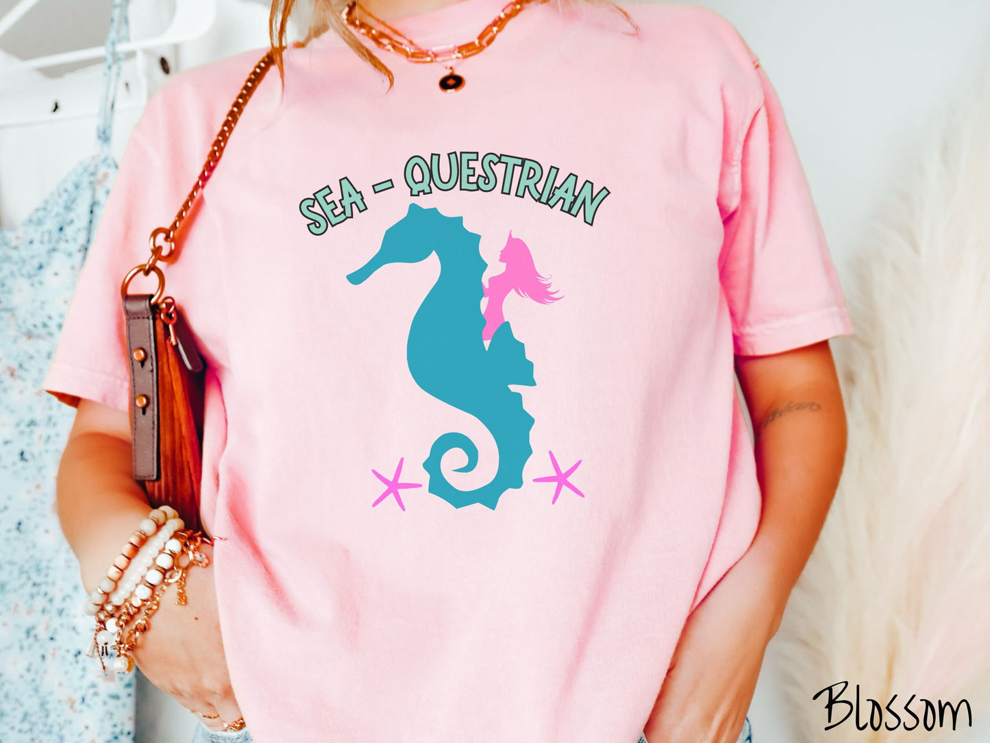 A woman wearing a cute, vintage blossom colored Comfort Colors shirt with text Sea-Questrian in seafoam colored font across the top. Below this is a pink mermaid with flowing hair riding on the back of a turquoise seahorse with pink starfish below.
