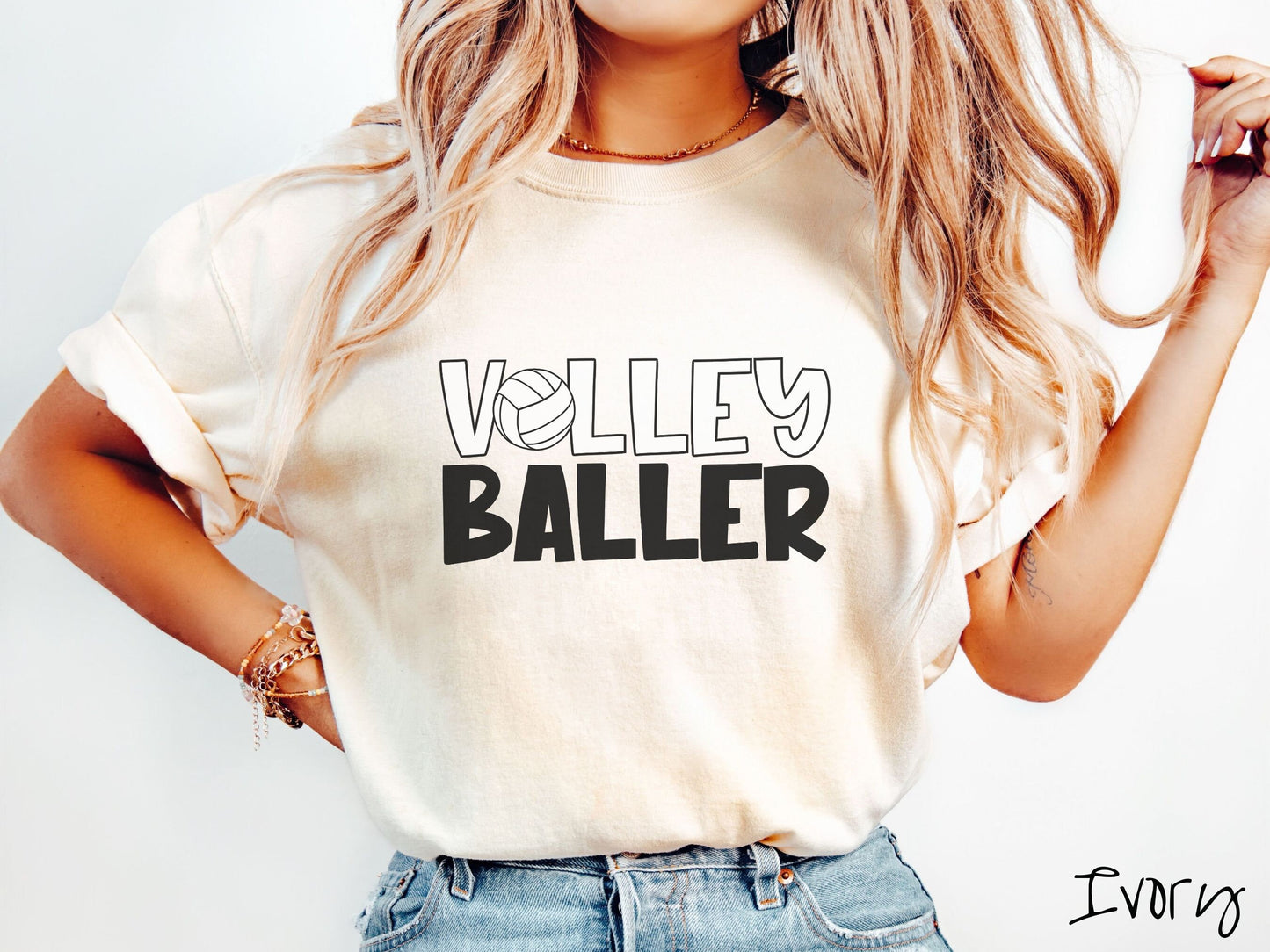 A woman wearing a cute, vintage ivory colored Comfort Colors shirt with the word Volley across the front in white, uppercase font. The O in Volley is a white volleyball. Below that is the word Baller in black, uppercase font.