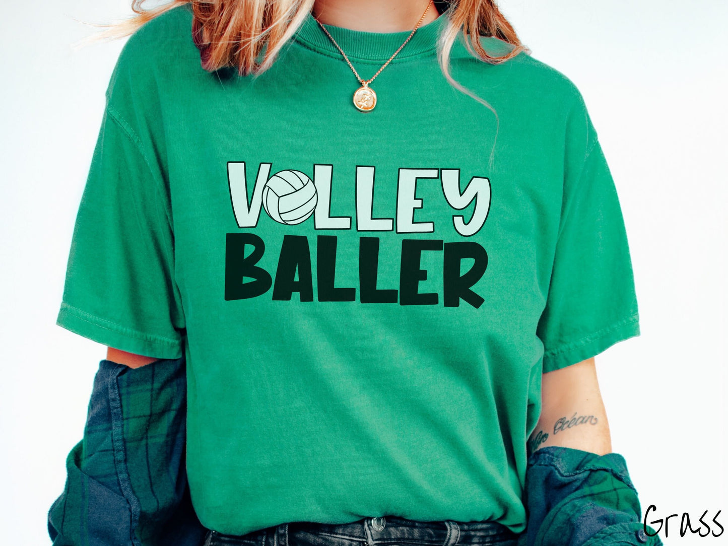 A woman wearing a cute, vintage grass colored Comfort Colors shirt with the word Volley across the front in white, uppercase font. The O in Volley is a white volleyball. Below that is the word Baller in black, uppercase font.
