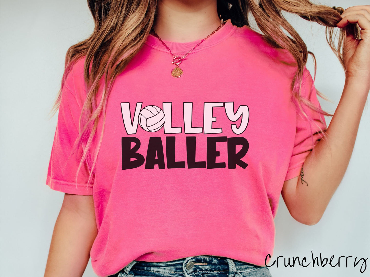 A woman wearing a cute, vintage crunchberry colored Comfort Colors shirt with the word Volley across the front in white, uppercase font. The O in Volley is a white volleyball. Below that is the word Baller in black, uppercase font.