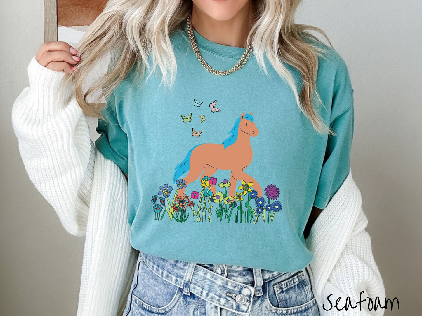 A woman wearing a cute, vintage seafoam colored Comfort Colors shirt with a brown horse with blue hair walking through a field of colorful flowers and grass. There are colorful butterflies flying above the horse.