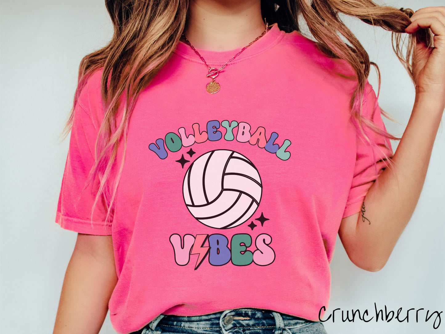 A woman wearing a cute, vintage crunchberry colored Comfort Colors shirt with the word Volleyball across the front in colorful, uppercase fonts. Below that is a white volleyball and below that is the word vibes in colorful, uppercase fonts.