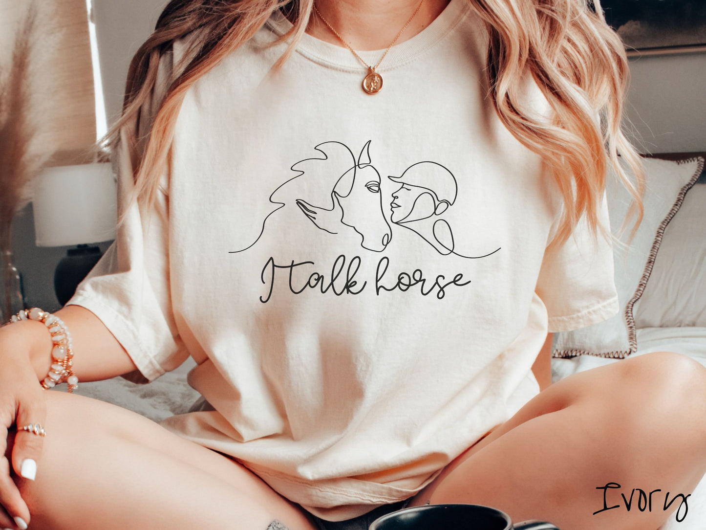 A woman wearing a cute, vintage ivory colored Comfort Colors shirt with a black silhouette outline of a horse jockey embracing his horse. Below that are the words I talk horse in black, cursive font.