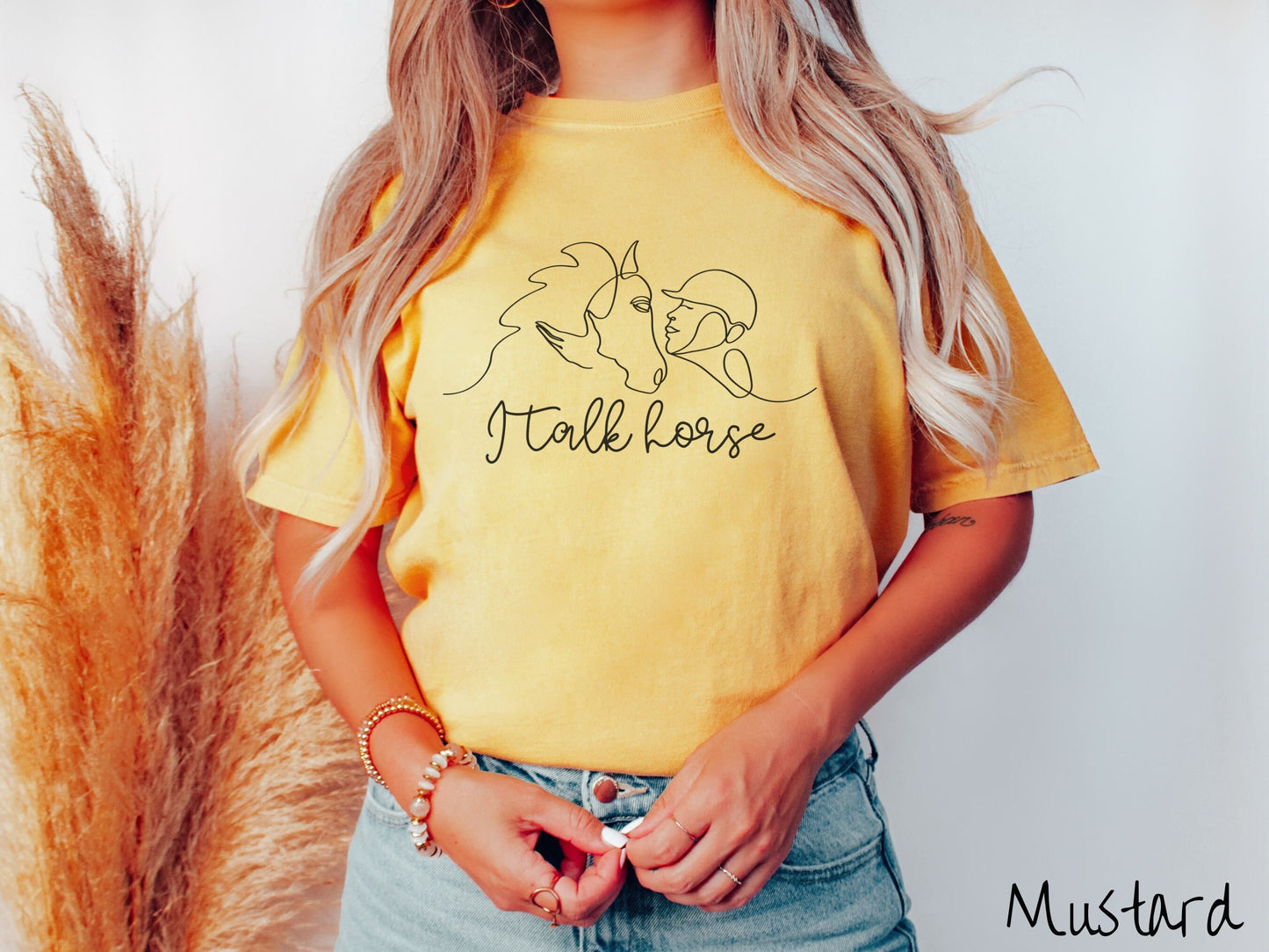 A woman wearing a cute, vintage mustard colored Comfort Colors shirt with a black silhouette outline of a horse jockey embracing his horse. Below that are the words I talk horse in black, cursive font.