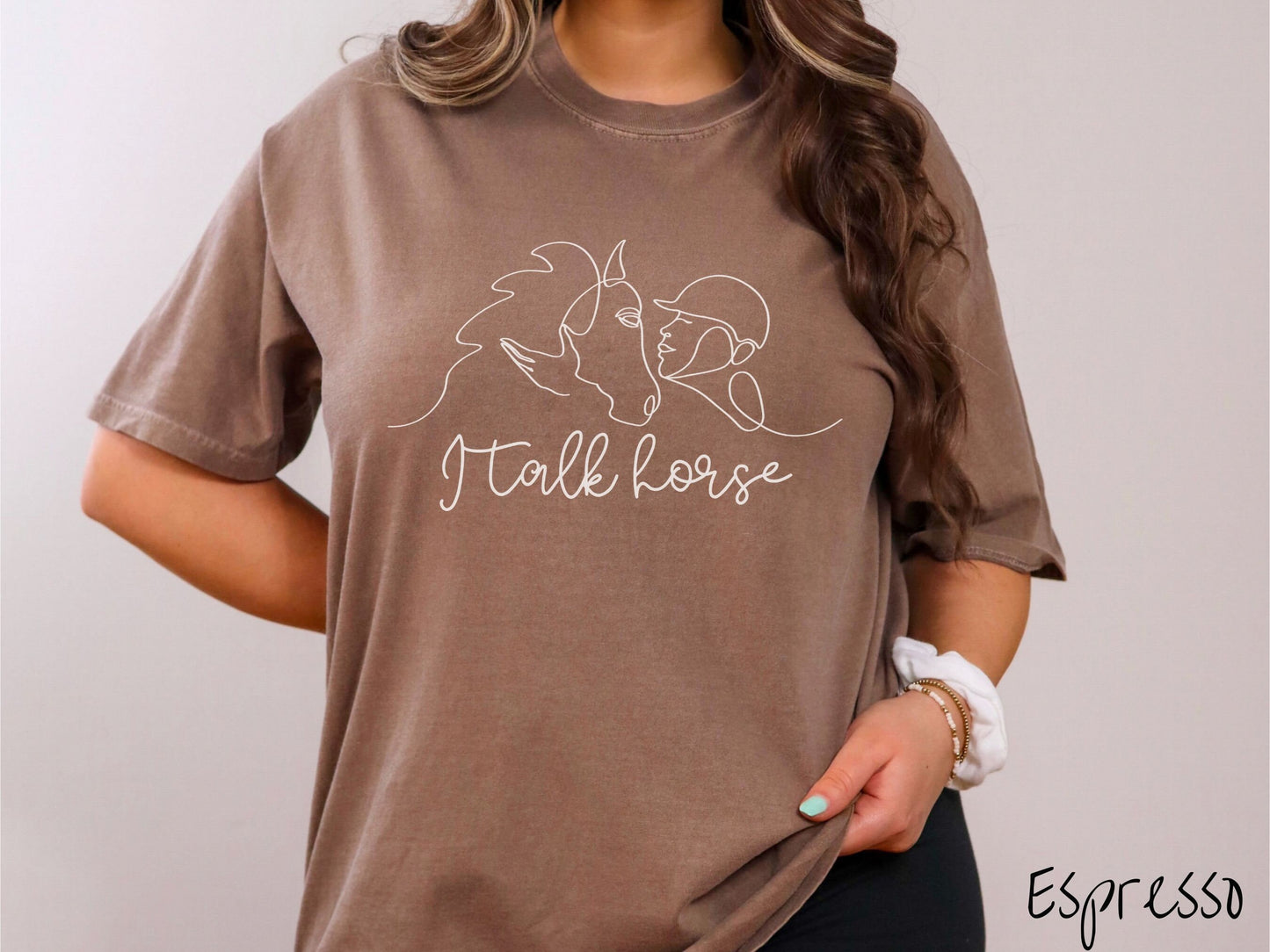 A woman wearing a cute, vintage espresso colored Comfort Colors shirt with a white silhouette outline of a horse jockey embracing his horse. Below that are the words I talk horse in white, cursive font.