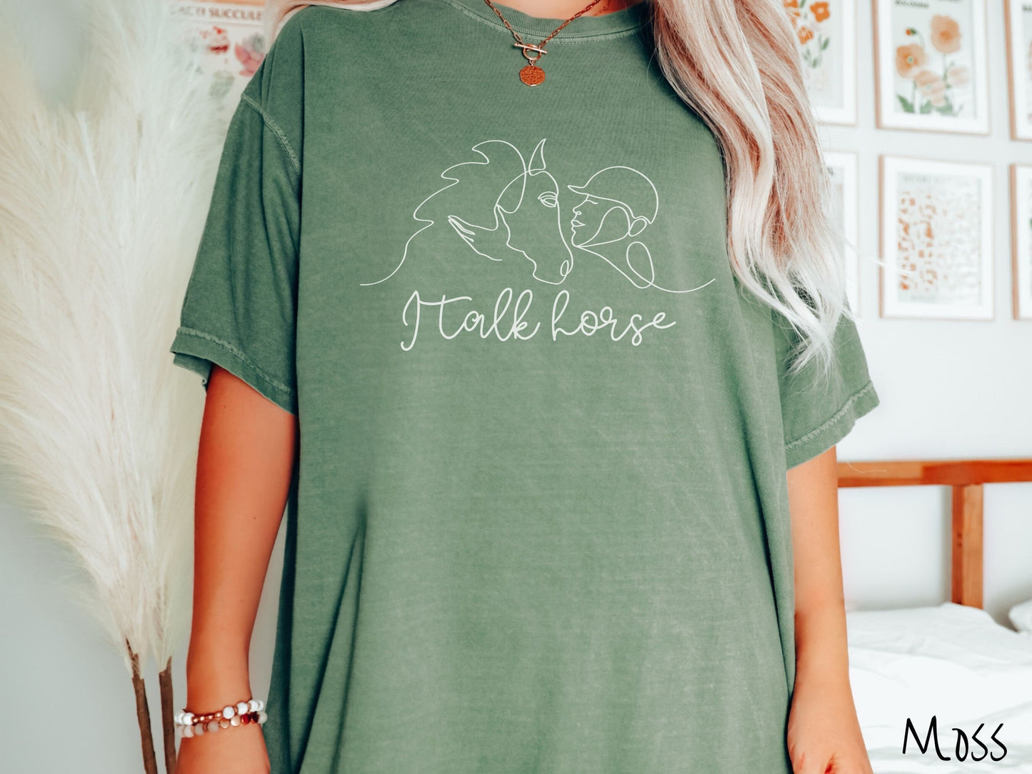 A woman wearing a cute, vintage moss colored Comfort Colors shirt with a white silhouette outline of a horse jockey embracing his horse. Below that are the words I talk horse in white, cursive font.