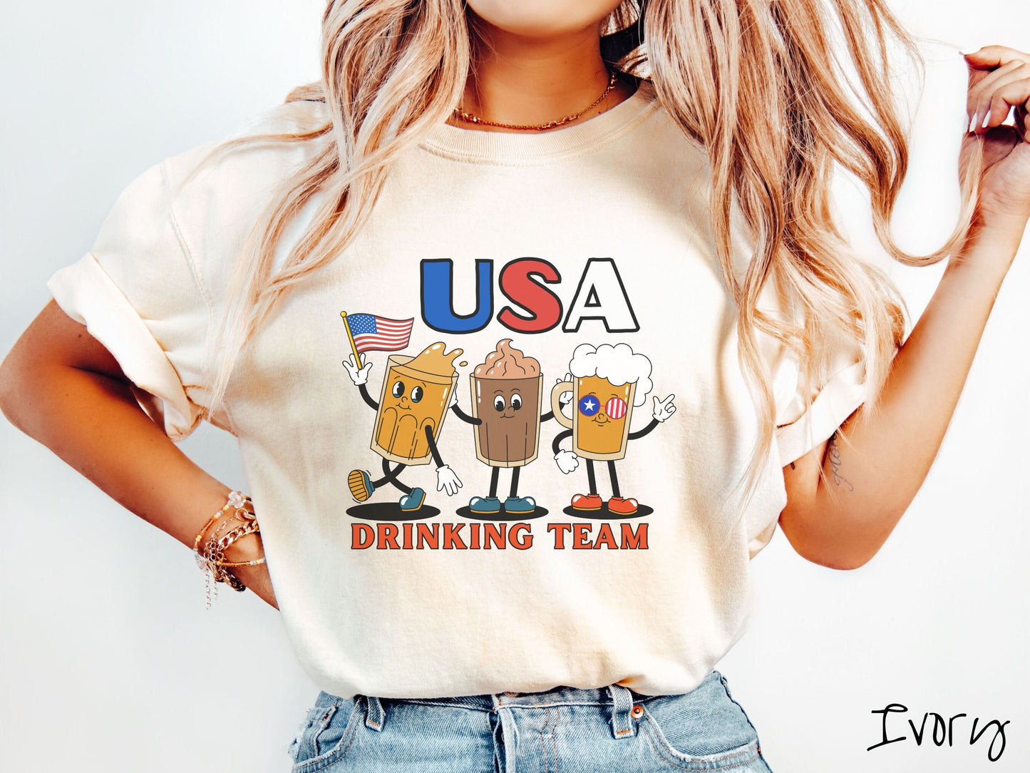 A woman wearing a cute, vintage ivory colored shirt with the letters USA in red, white and blue font across the top. Below are three mugs of beer with hands and feet holding American flags, below them is the text Drinking Team in red font.