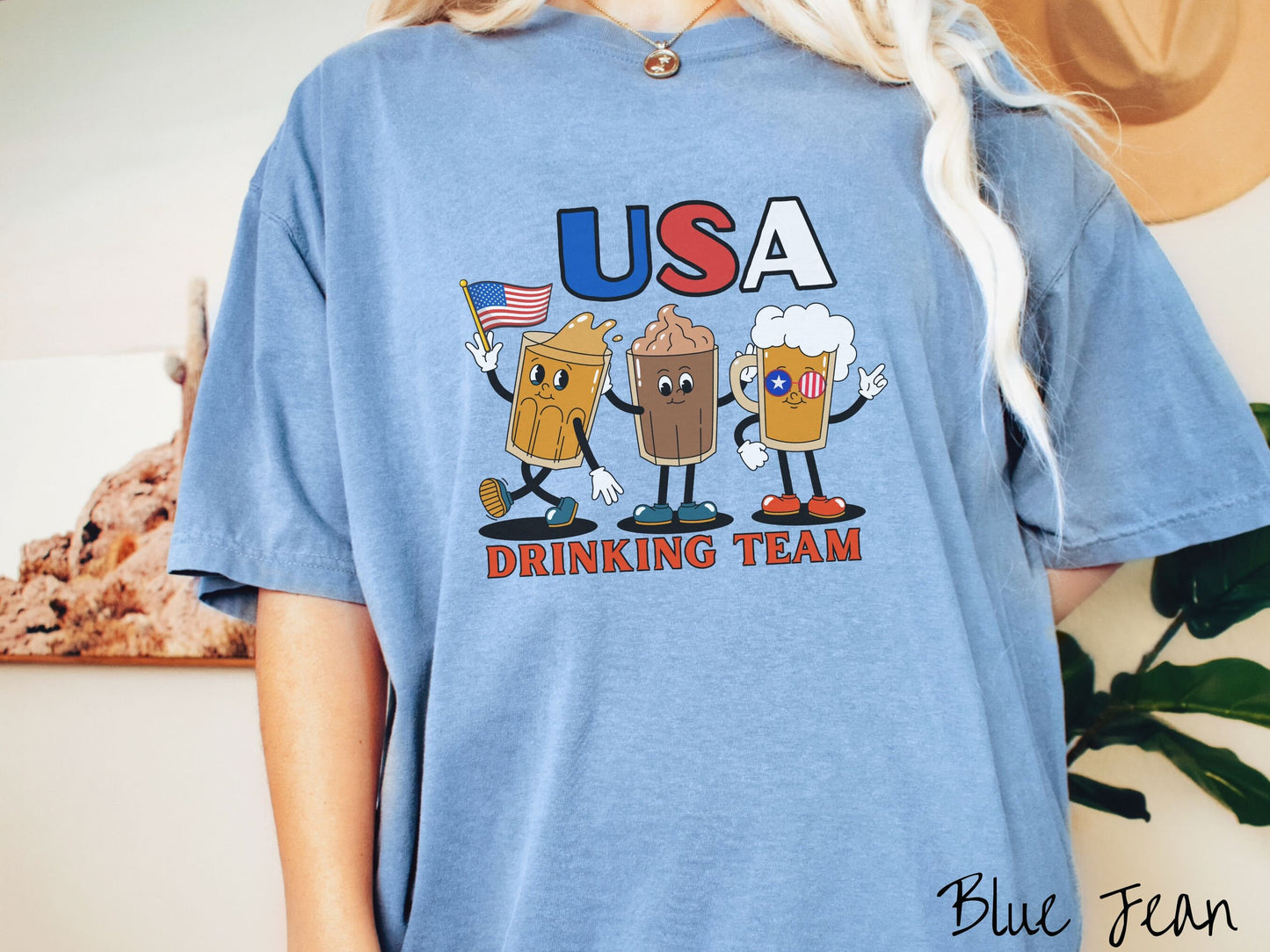 A woman wearing a cute, vintage blue jean colored shirt with the letters USA in red, white and blue font across the top. Below are three mugs of beer with hands and feet holding American flags, below them is the text Drinking Team in red font.
