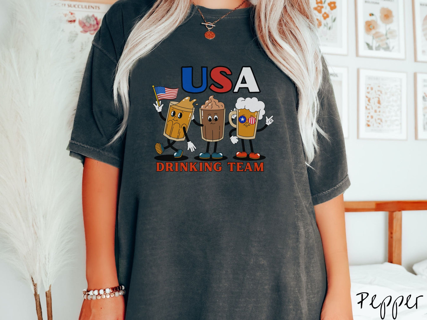 A woman wearing a cute, vintage pepper colored shirt with the letters USA in red, white and blue font across the top. Below are three mugs of beer with hands and feet holding American flags, below them is the text Drinking Team in red font.