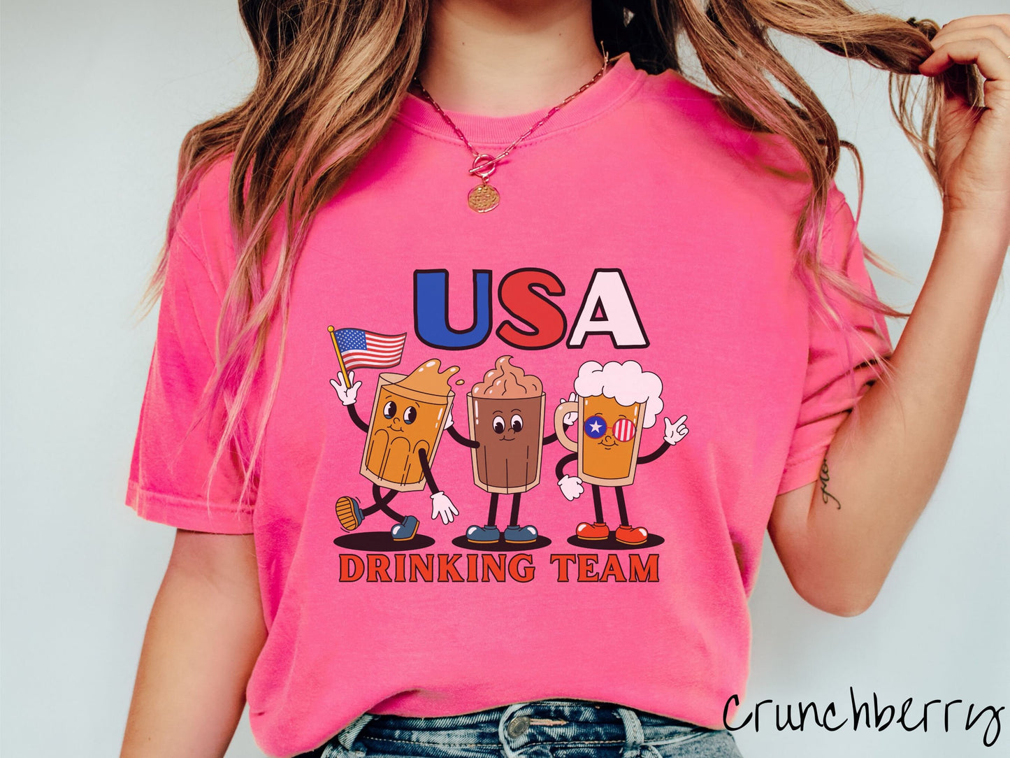 A woman wearing a cute, vintage crunchberry colored shirt with the letters USA in red, white and blue font across the top. Below are three mugs of beer with hands and feet holding American flags, below them is the text Drinking Team in red font.