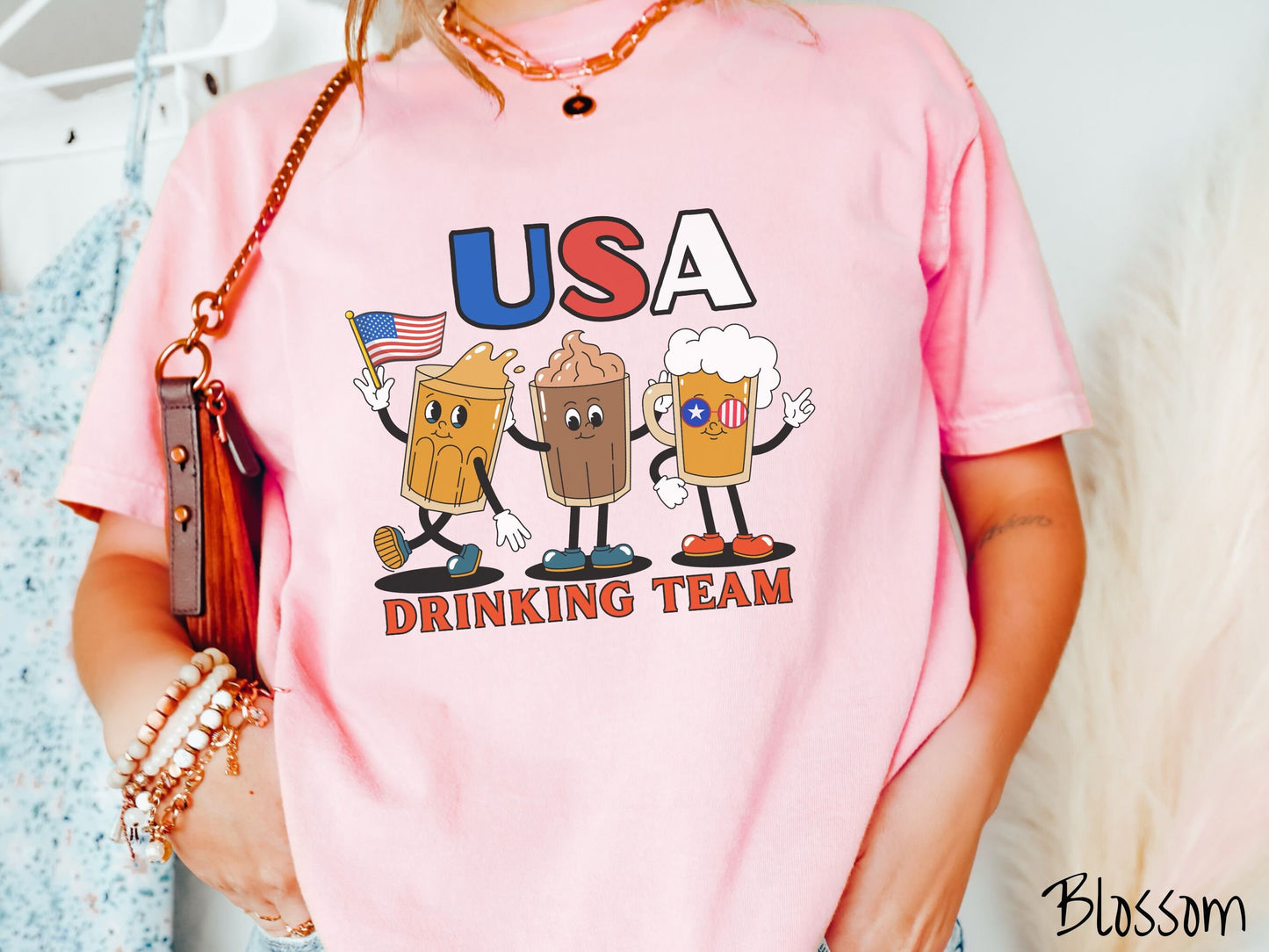 A woman wearing a cute, vintage blossom colored shirt with the letters USA in red, white and blue font across the top. Below are three mugs of beer with hands and feet holding American flags, below them is the text Drinking Team in red font.