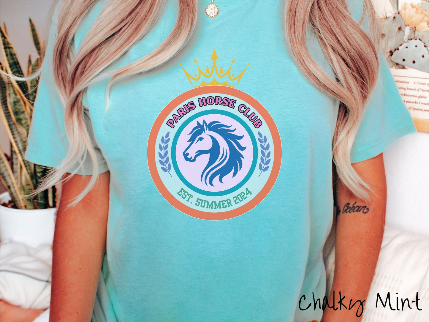 A woman wearing a cute, vintage chalky mint colored Comfort Colors shirt with a colorful coat of arms with a horse in the middle of two concentric circles, a crown on top. There is text along the top Paris Horse Club and below that Est. Summer 2024.