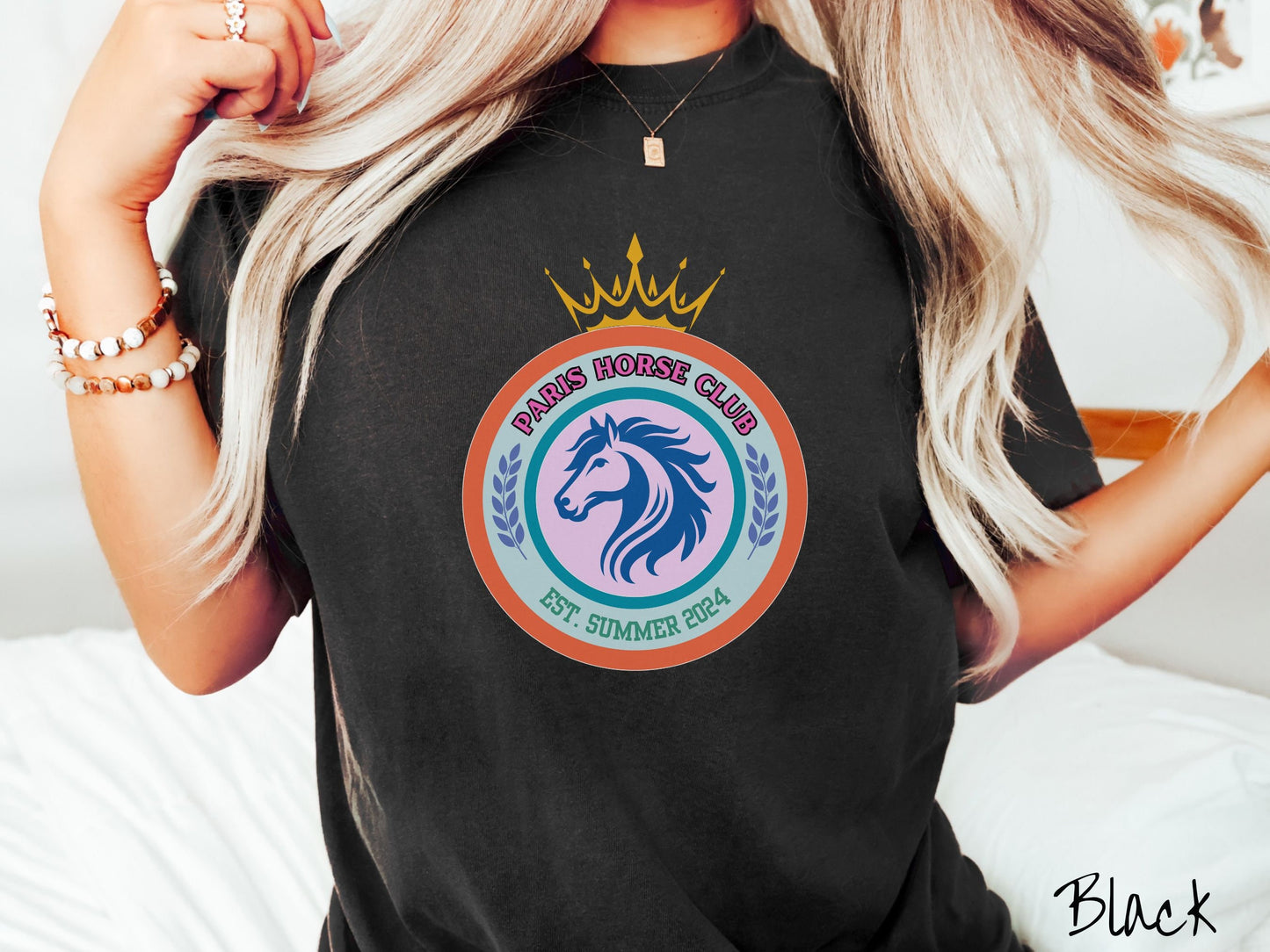A woman wearing a cute, vintage black colored Comfort Colors shirt with a colorful coat of arms with a horse in the middle of two concentric circles, a crown on top. There is pink text along the top Paris Horse Club and below that Est. Summer 2024.