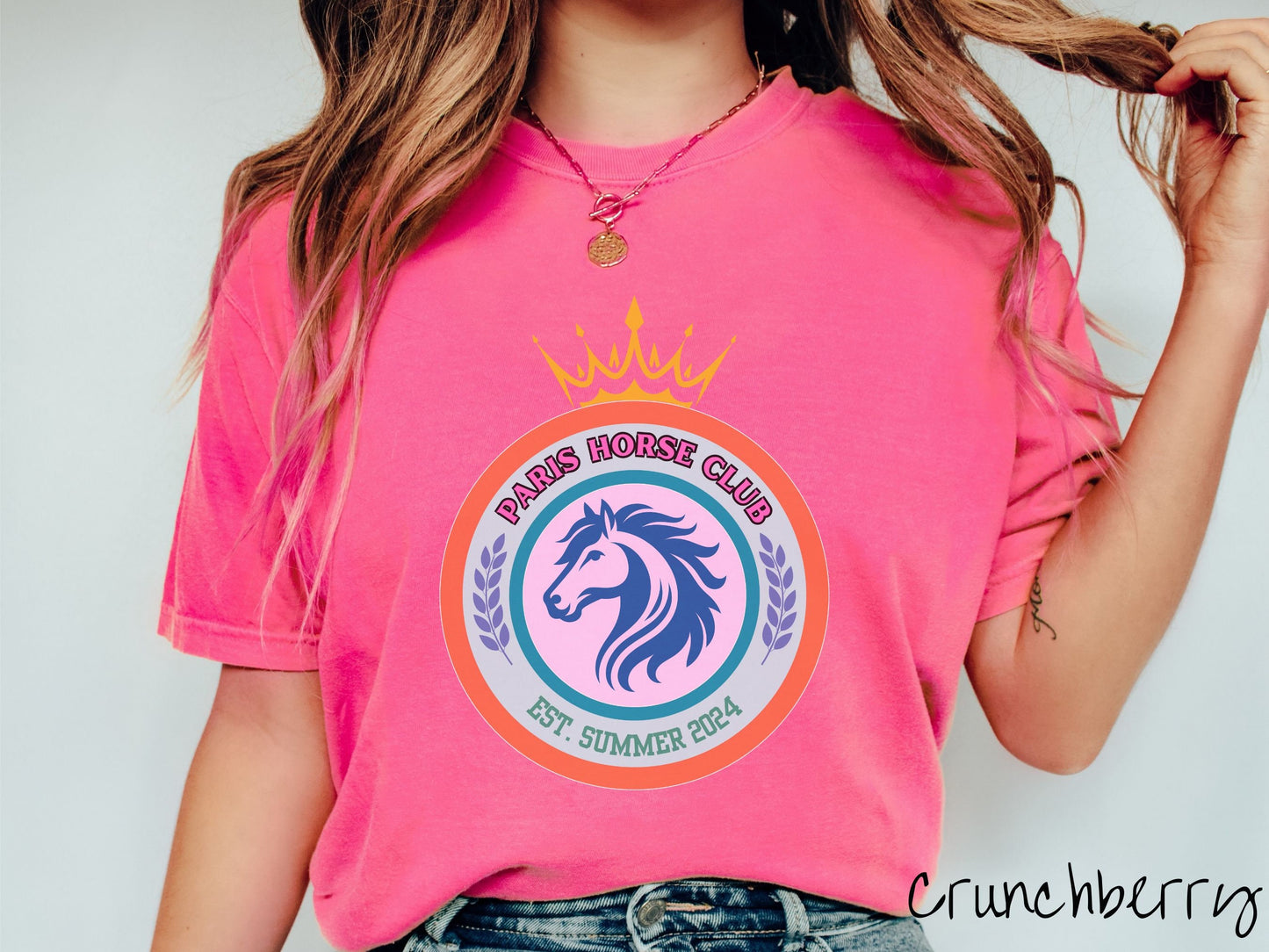 A woman wearing a cute, vintage crunchberry colored Comfort Colors shirt with a colorful coat of arms with a horse in the middle of two concentric circles, a crown on top. There is text along the top Paris Horse Club and below that Est. Summer 2024.