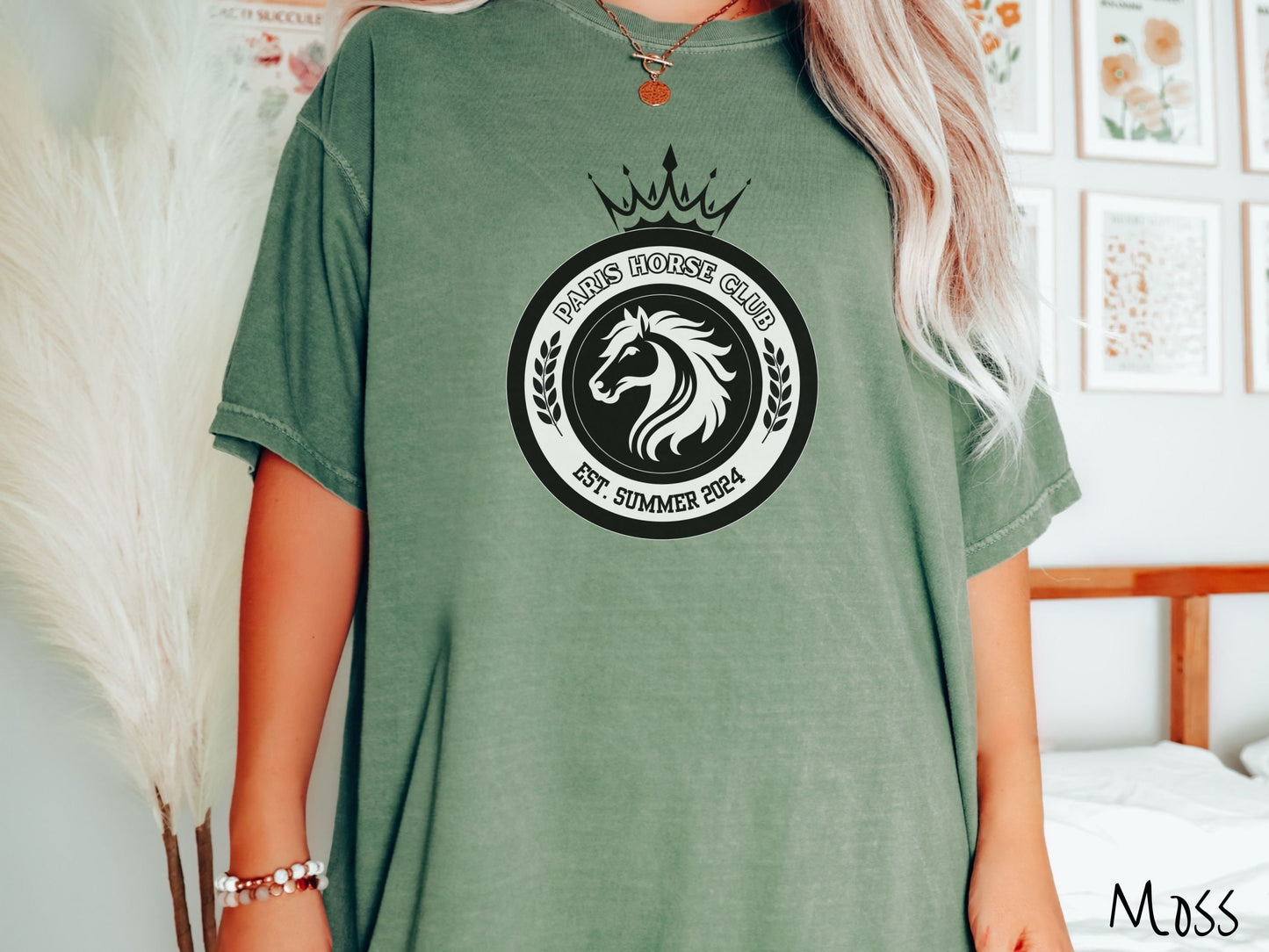 A woman wearing a cute, vintage moss colored Comfort Colors shirt with a black and white coat of arms with a horse in the middle of two concentric circles, a crown on top. There is text along the top Paris Horse Club and below that Est. Summer 2024.