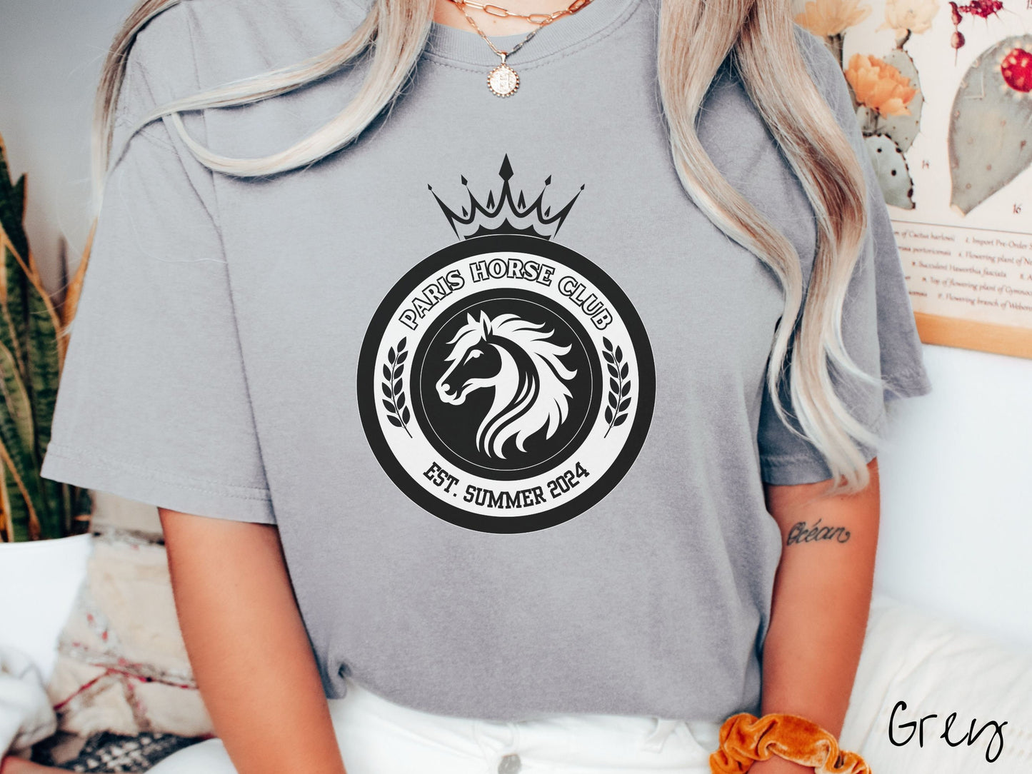 A woman wearing a cute, vintage grey colored Comfort Colors shirt with a black and white coat of arms with a horse in the middle of two concentric circles, a crown on top. There is text along the top Paris Horse Club and below that Est. Summer 2024.