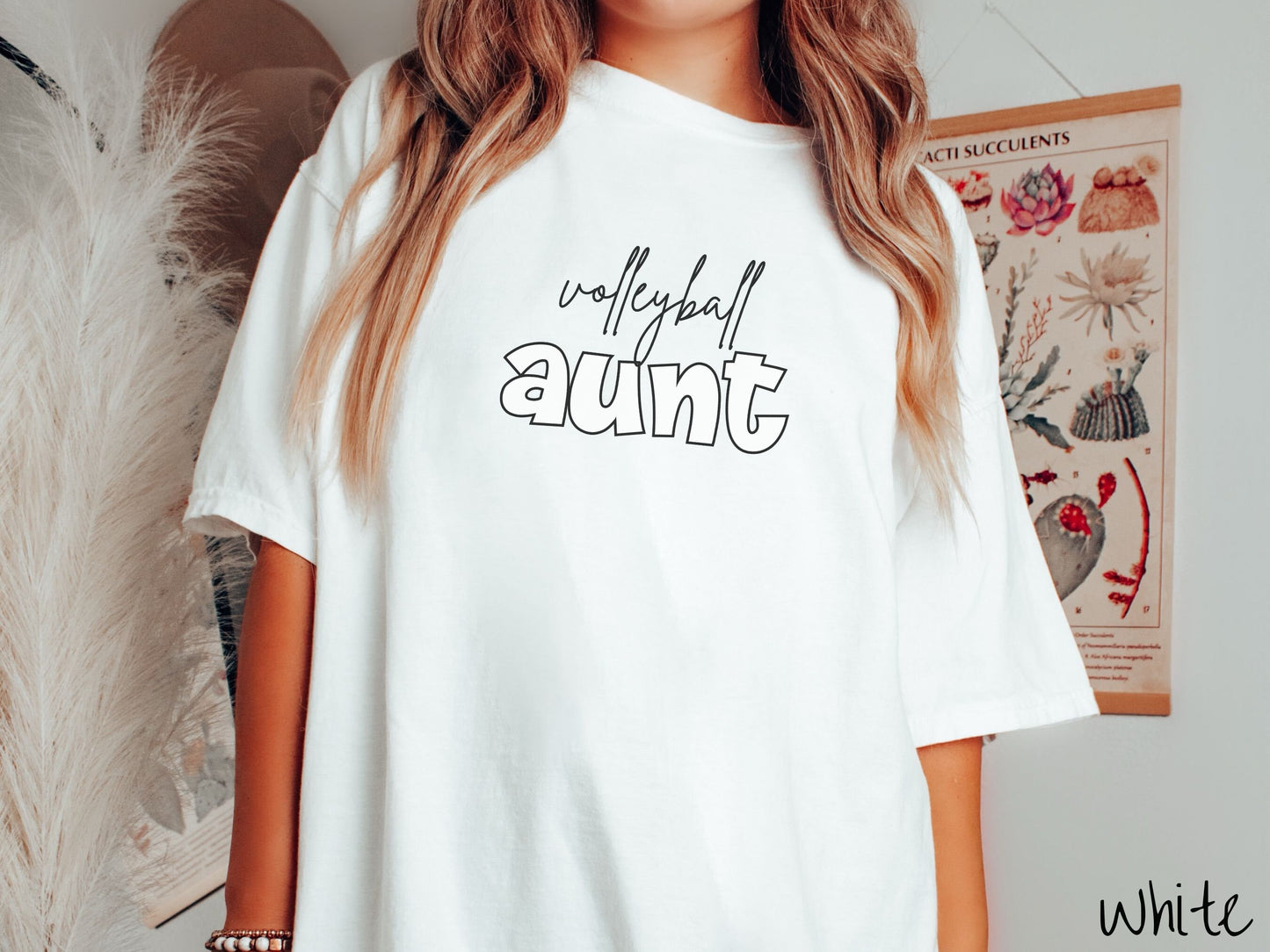 A woman wearing a cute, vintage white colored Comfort Colors shirt with the word volleyball across the front in black, undercase cursive writing. Below that is the word aunt in large, white font.