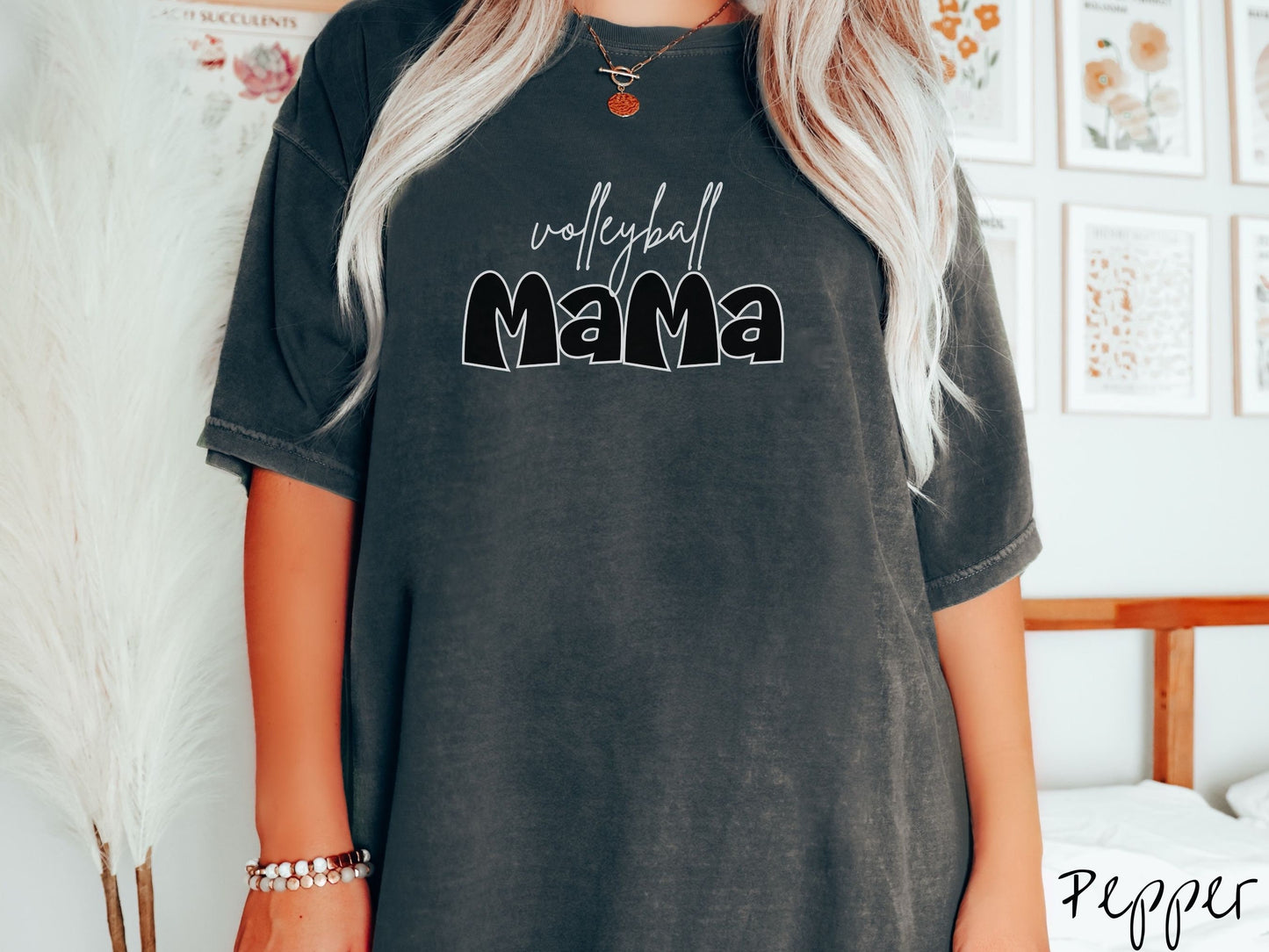 A woman wearing a cute, vintage pepper colored Comfort Colors shirt with the word volleyball across the front in white, undercase cursive writing. Below that is the word Mama in large, black font.