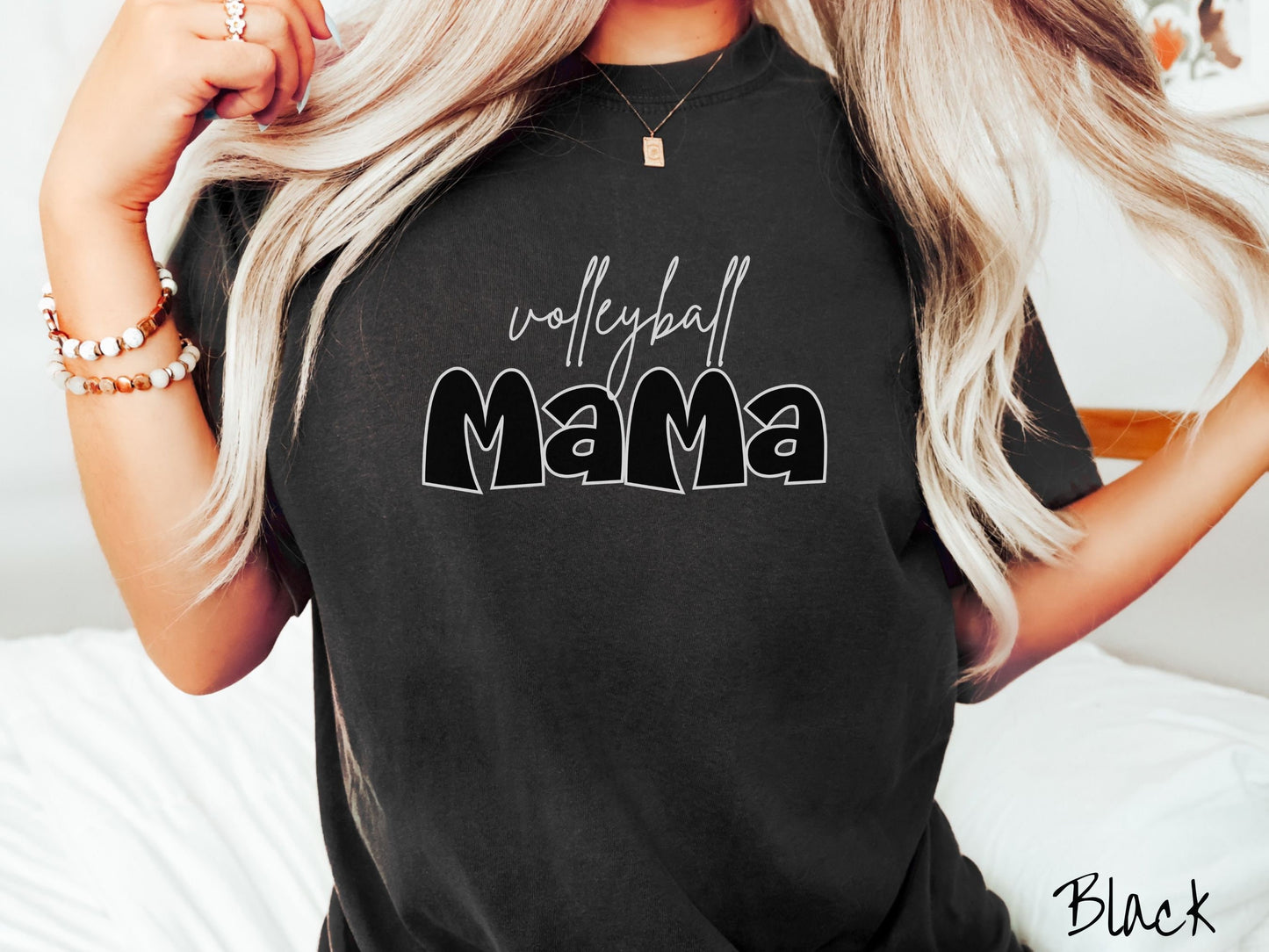 A woman wearing a cute, vintage black colored Comfort Colors shirt with the word volleyball across the front in white, undercase cursive writing. Below that is the word Mama in large, black font.