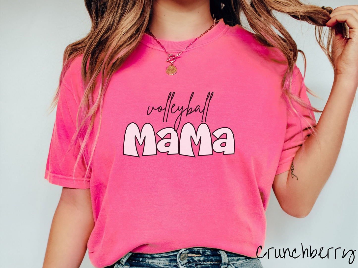 A woman wearing a cute, vintage crunchberry colored Comfort Colors shirt with the word volleyball across the front in black, undercase cursive writing. Below that is the word Mama in large, light pink font.