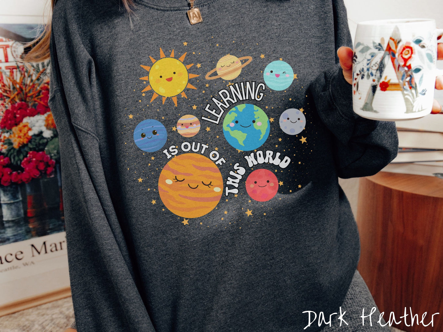 A woman wearing a cute, vintage dark heather colored sweatshirt showing the solar system and the planets have smiling faces. There is text saying Learning Is Out of This World among the planets, and there are many stars twinkling in the background.