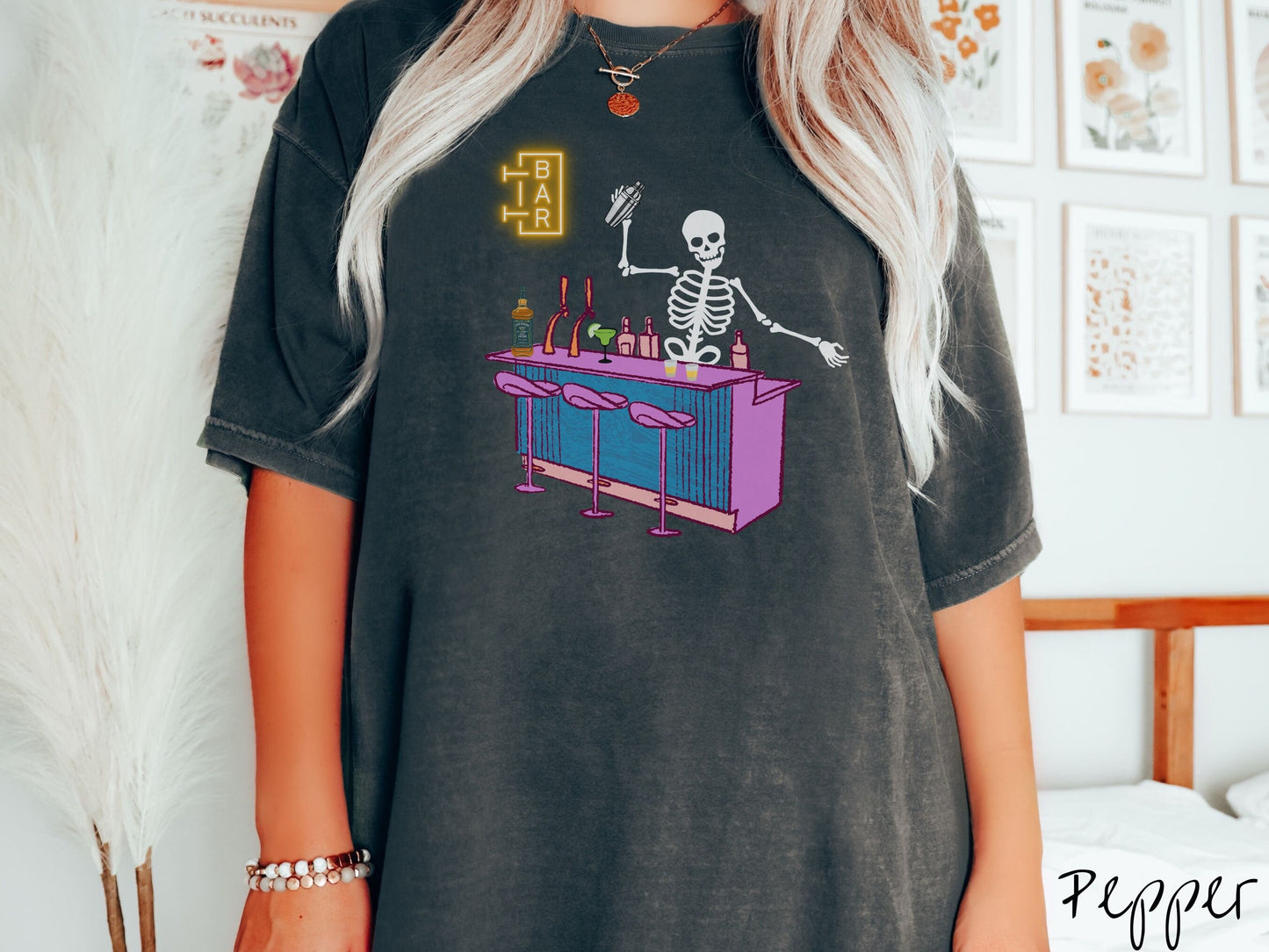 A woman wearing a cute, vintage pepper colored Comfort Colors t-shirt showing a skeleton bartender behind a purple bar mixing drinks. There is a yellow bar sign behind the bar and three purple chairs in front. There are liquor bottles on top.