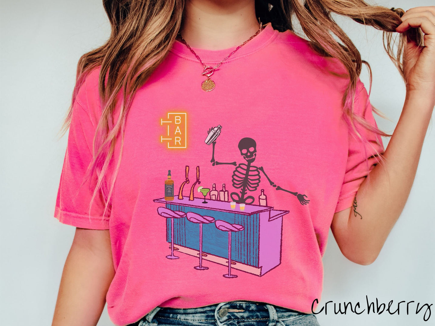 A woman wearing a cute, vintage crunchberry colored Comfort Colors t-shirt showing a skeleton bartender behind a purple bar mixing drinks. There is a yellow bar sign behind the bar and three purple chairs in front. There are liquor bottles on top.