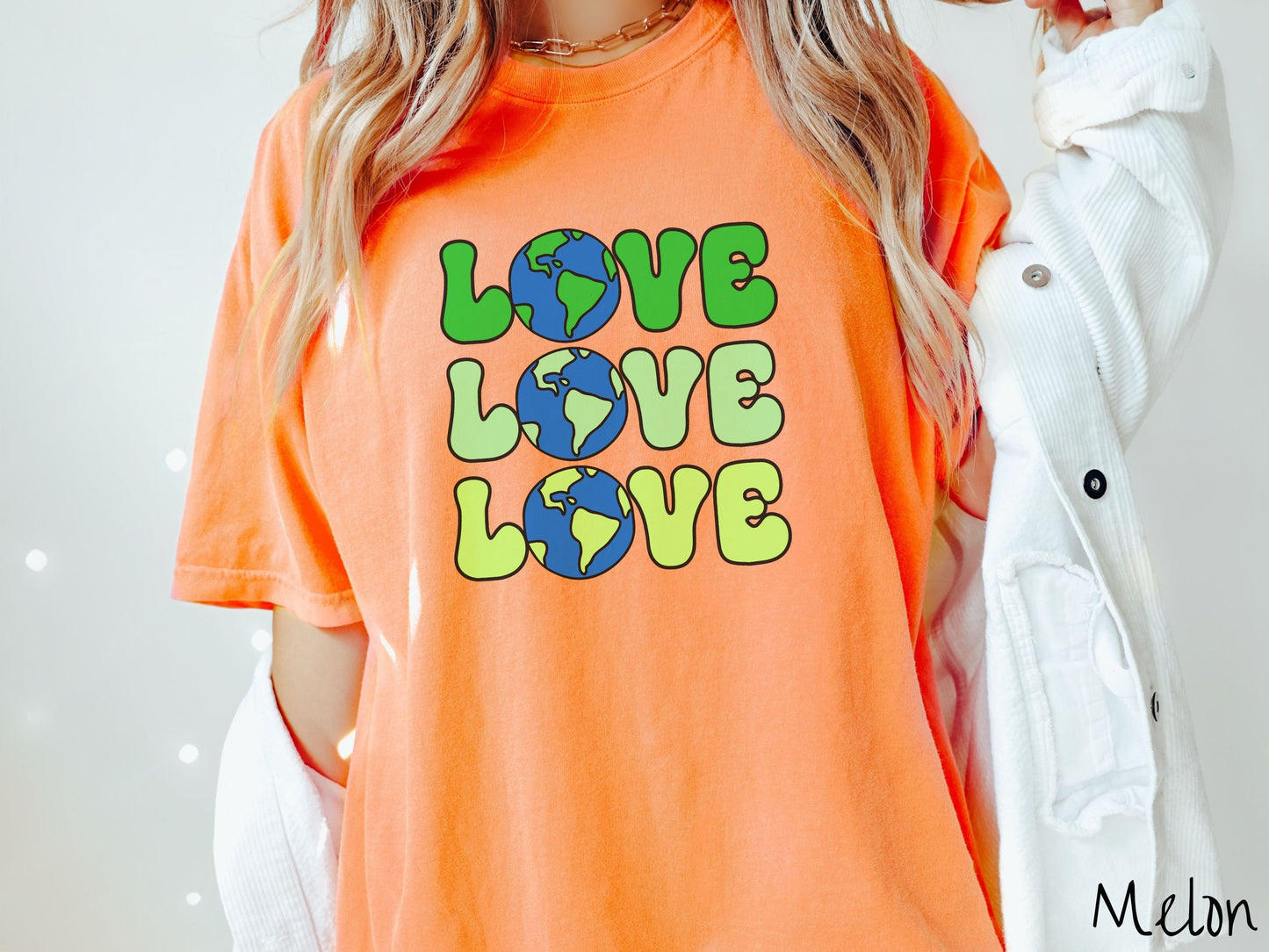 A woman wearing a cute, vintage melon colored Comfort Colors shirt with the word Love listed vertically three times in varying shades of green, uppercase font. The Os in the words are pictures of the blue and green earth.