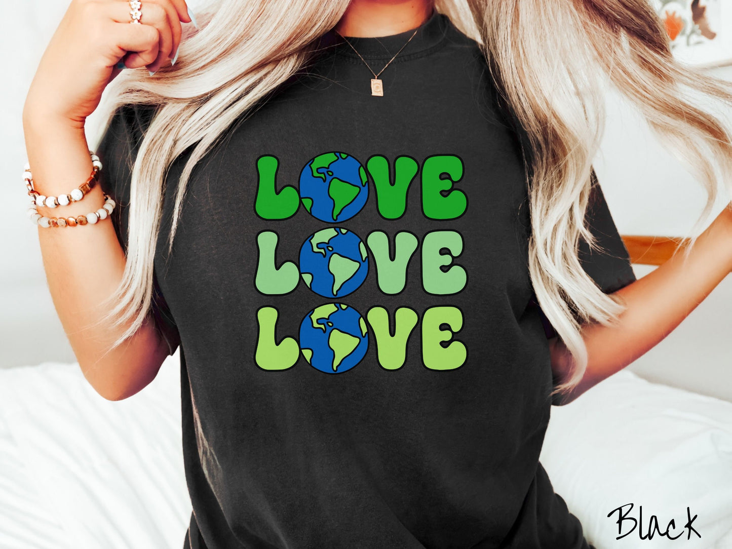 A woman wearing a cute, vintage black colored Comfort Colors shirt with the word Love listed vertically three times in varying shades of green, uppercase font. The Os in the words are pictures of the blue and green earth.
