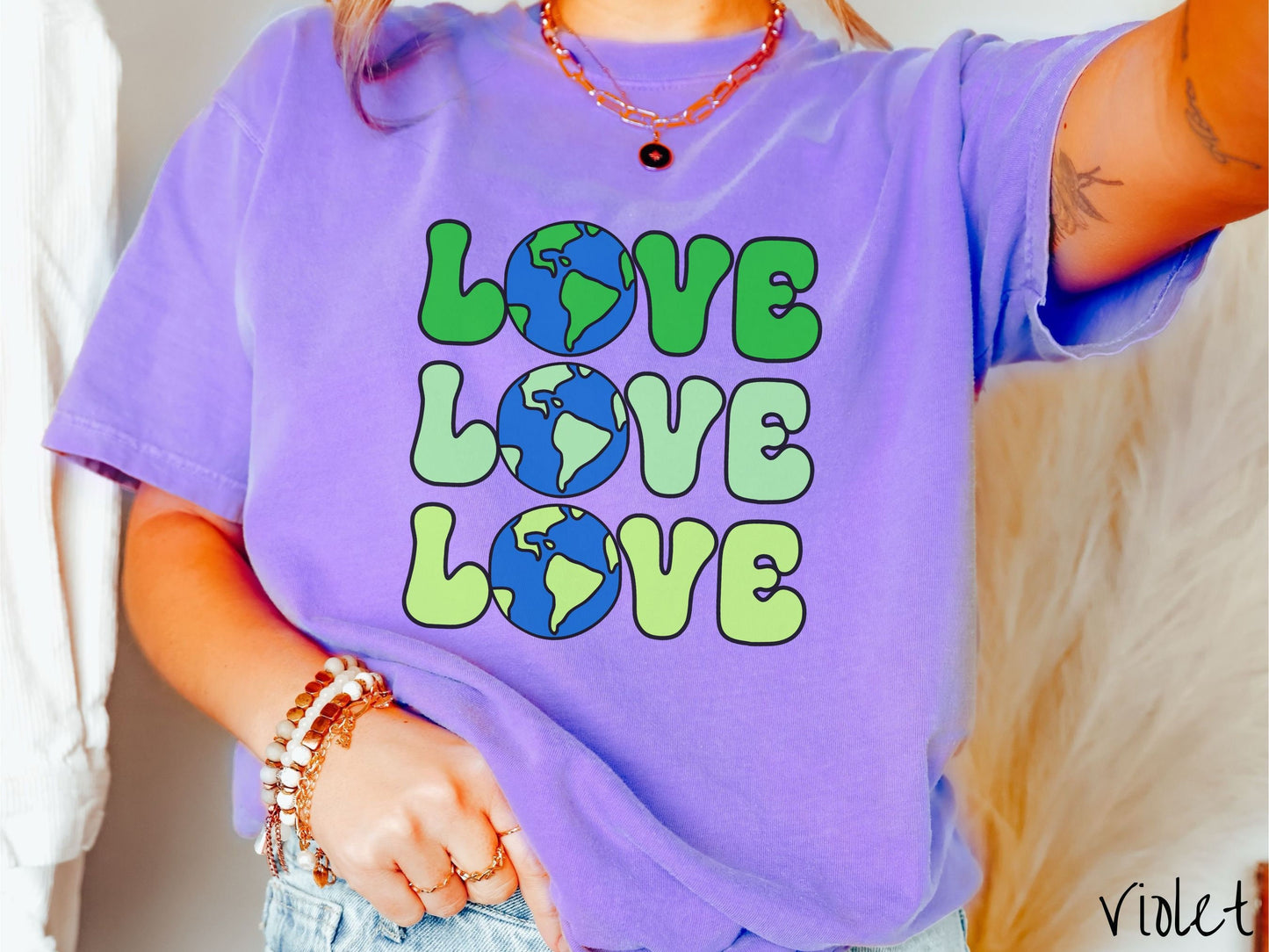 A woman wearing a cute, vintage violet colored Comfort Colors shirt with the word Love listed vertically three times in varying shades of green, uppercase font. The Os in the words are pictures of the blue and green earth.