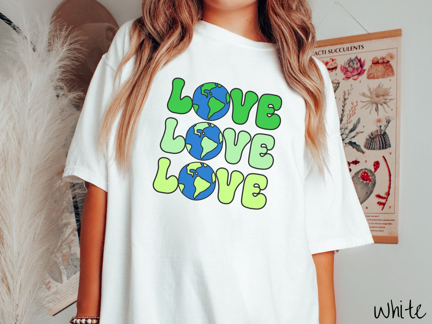 A woman wearing a cute, vintage white colored Comfort Colors shirt with the word Love listed vertically three times in varying shades of green, uppercase font. The Os in the words are pictures of the blue and green earth.