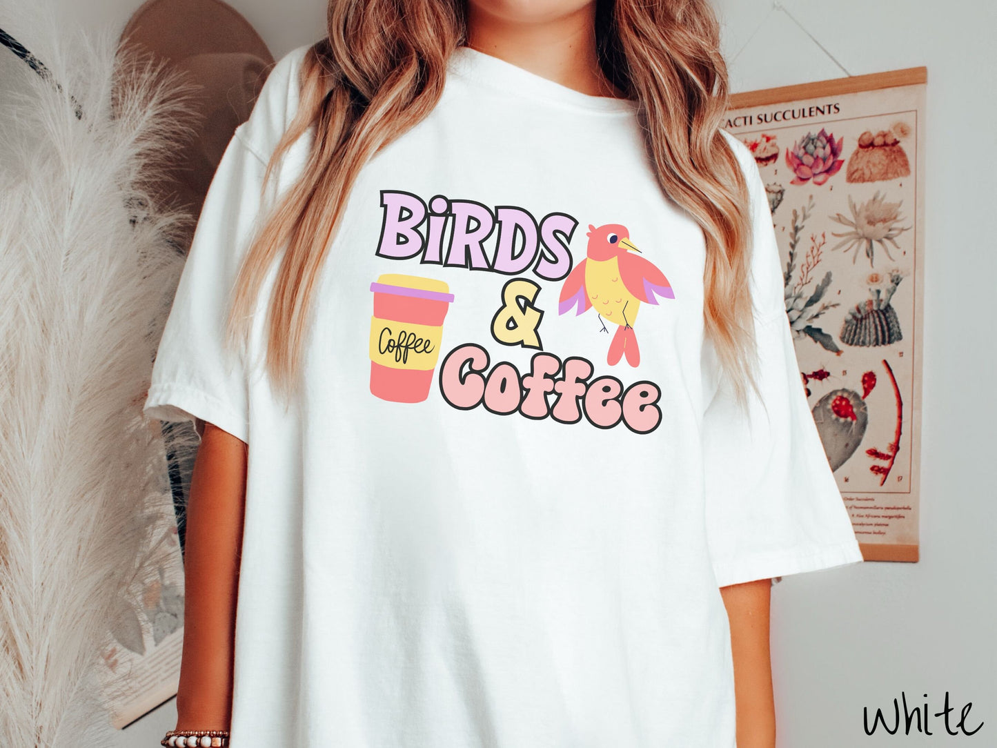 A woman wearing a cute, vintage white colored Comfort Colors shirt with the text Birds & Coffee in purple and light pink font. Next to the text is a red, yellow, and purple smiling bird in the air and a red and yellow to-go coffee cup.